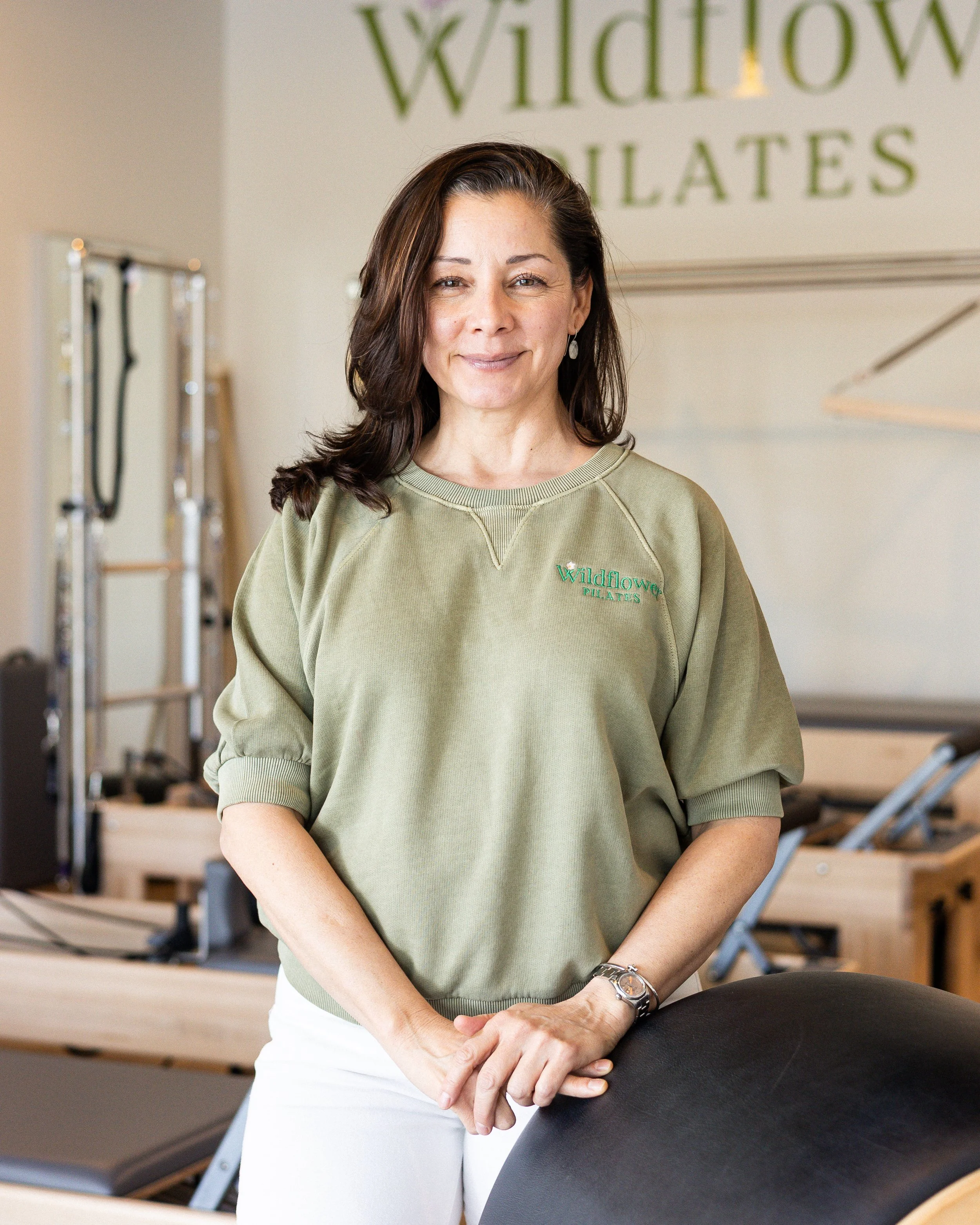 A woman with brown hair wearing a green sweatshirt with 'Wildflower Pilates' embroidered on it, standing in a Pilates studio, smiling at the camera.