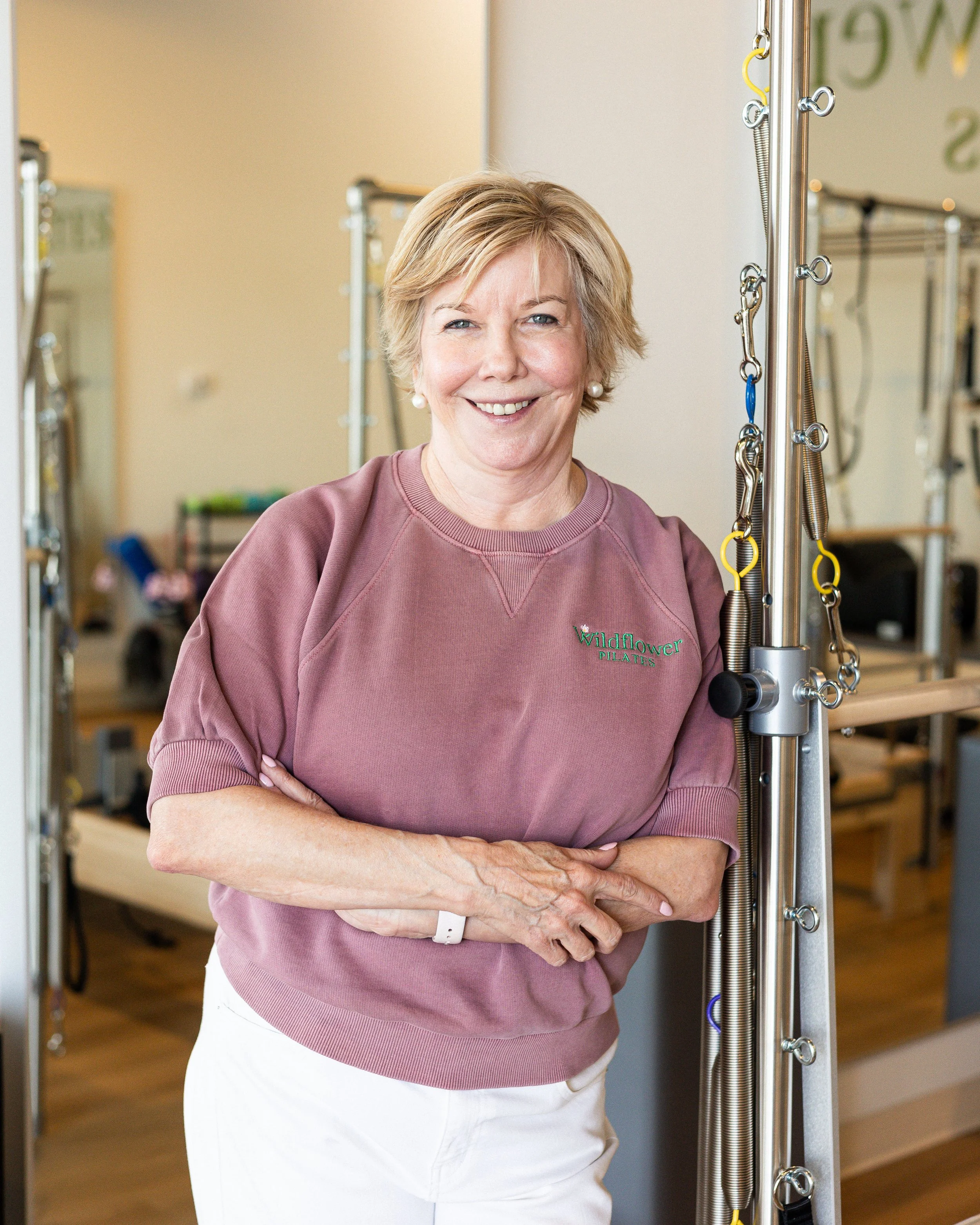 A smiling woman wearing a mauve sweatshirt with 'Wildflower Pilates' embroidered on it, standing with her arms crossed inside a Pilates studio.