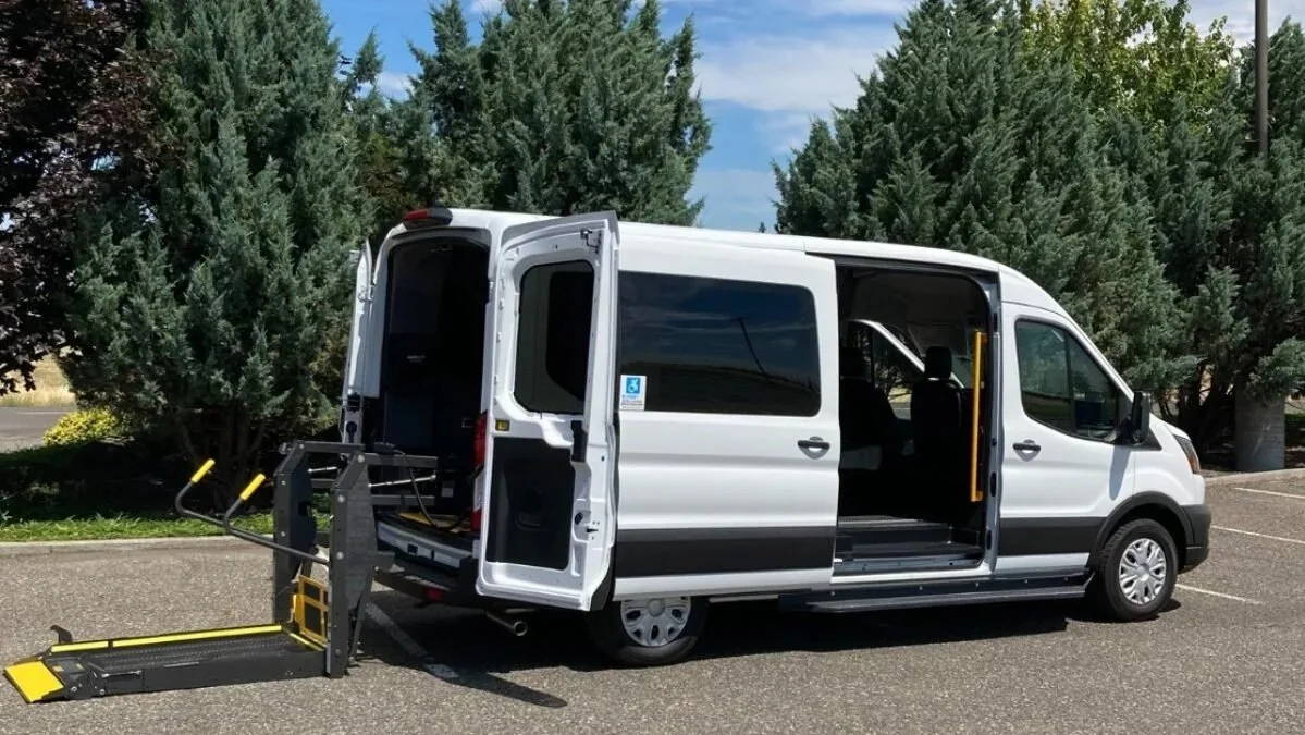 White passenger van with a side door open, showing a wheelchair lift ramp extended outside. The van is parked in an empty parking lot with tall green trees in the background.