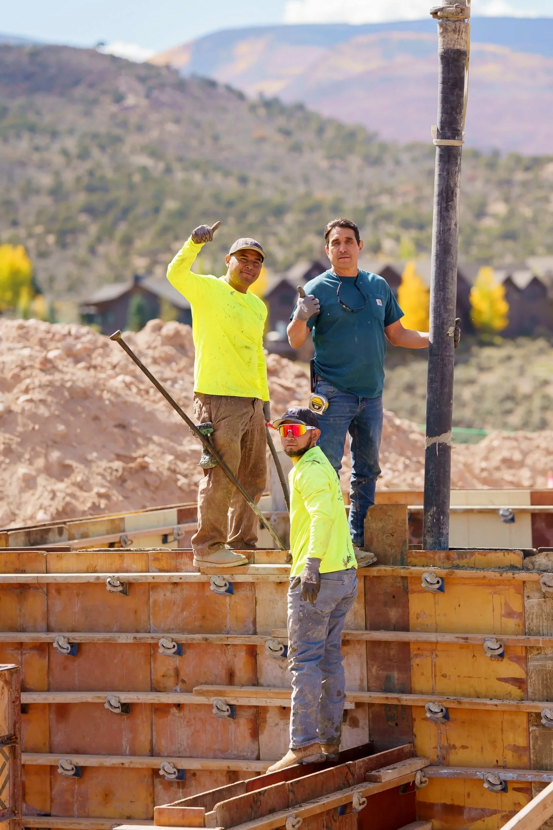 Three construction workers standing on a building foundation during daytime, with mountains and trees in the background. One worker is giving a thumbs-up and another is holding a tool, all wearing safety gear.
