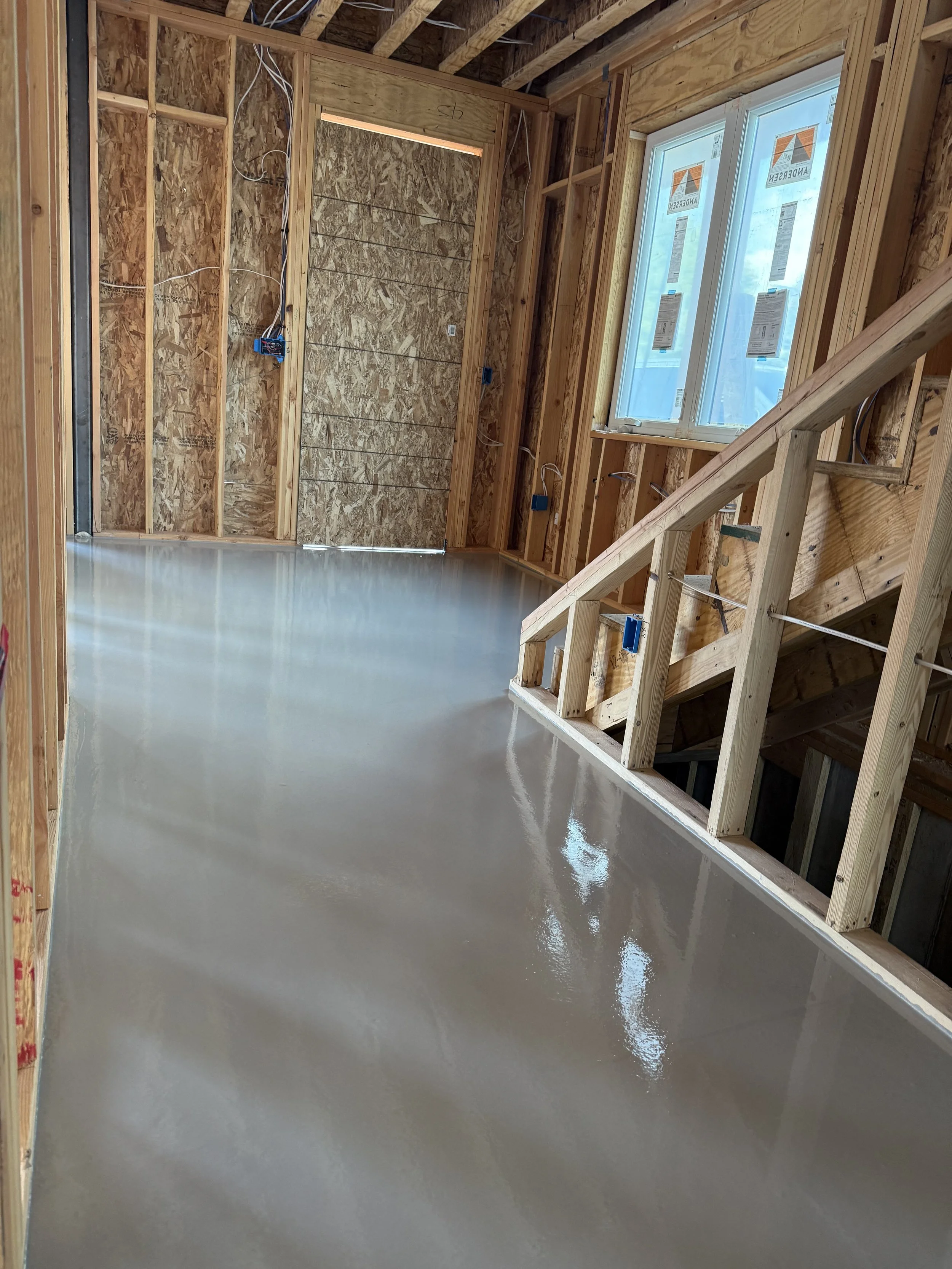 Interior of a house under construction with exposed wooden framing, electrical wiring, a window with protective coverings, and a freshly poured, shiny concrete floor.