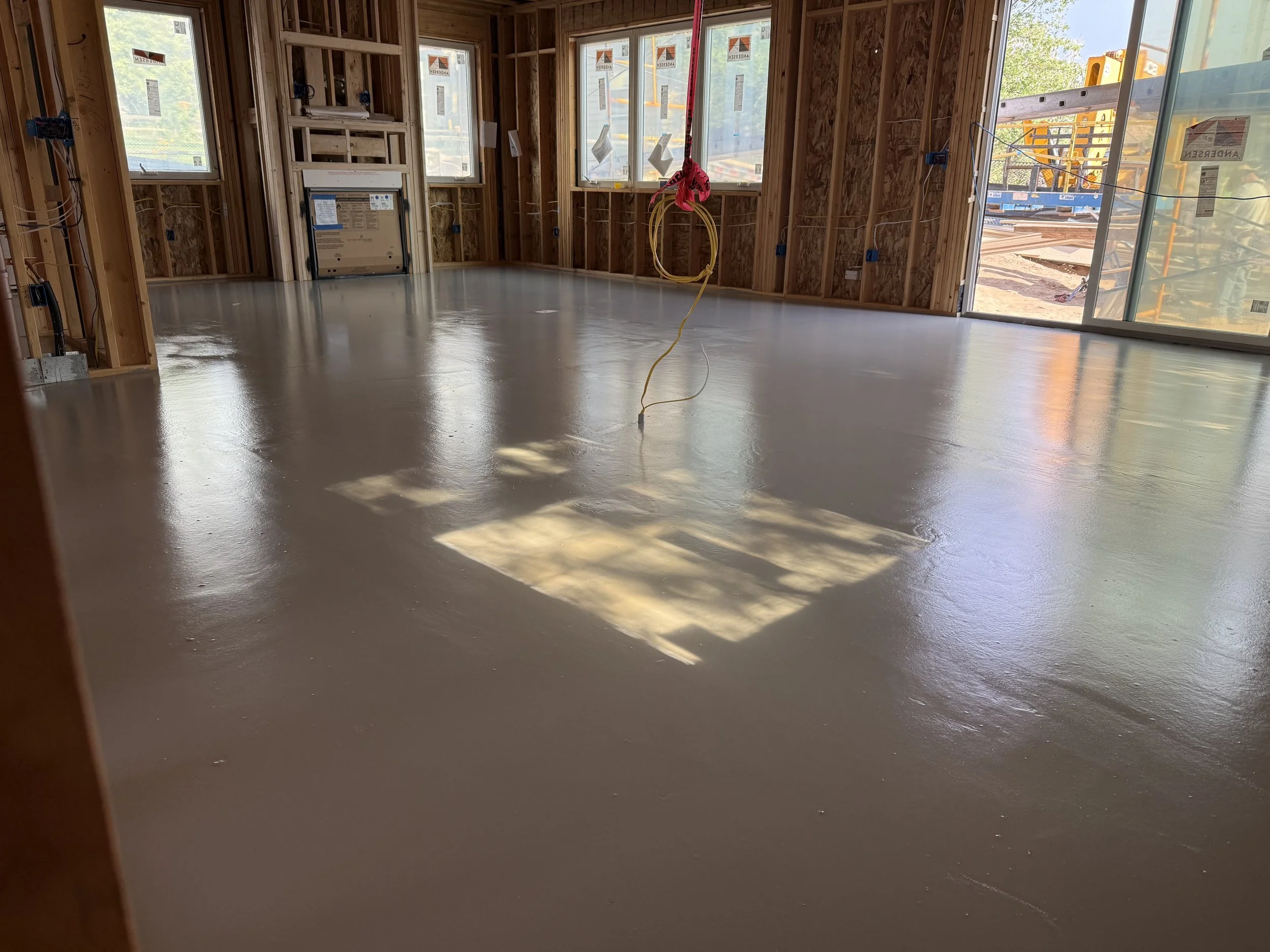 Interior view of a house under construction with a freshly poured concrete floor, open wooden wall studs, and large windows letting in natural light.