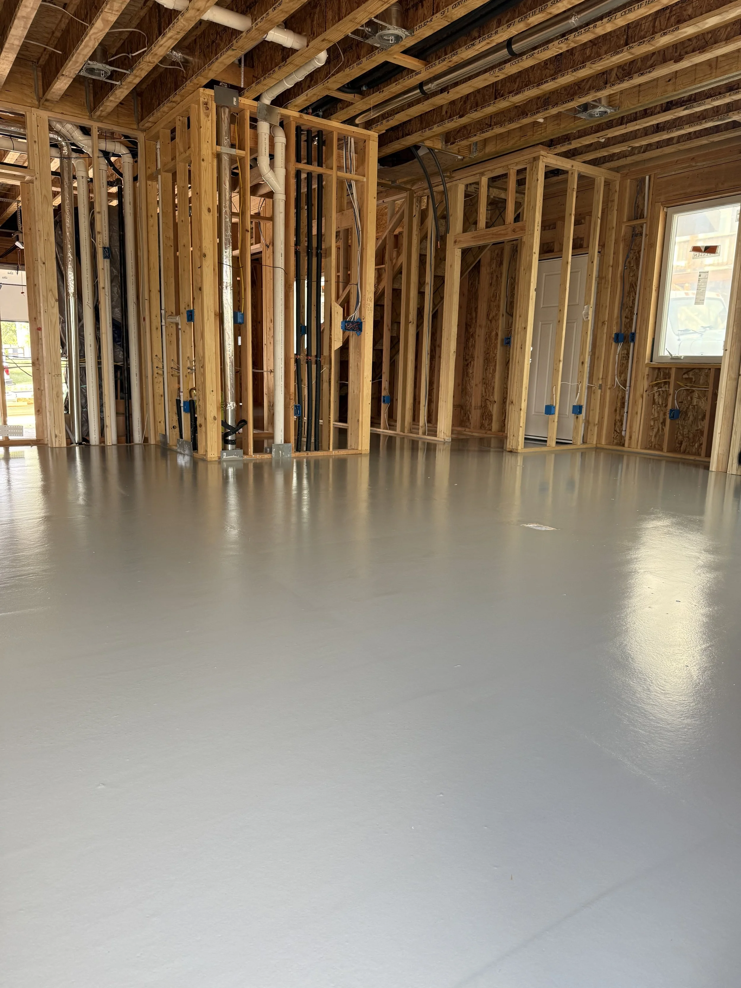 Interior view of a house under construction with exposed wooden framing, electrical wiring, and a freshly painted concrete floor.