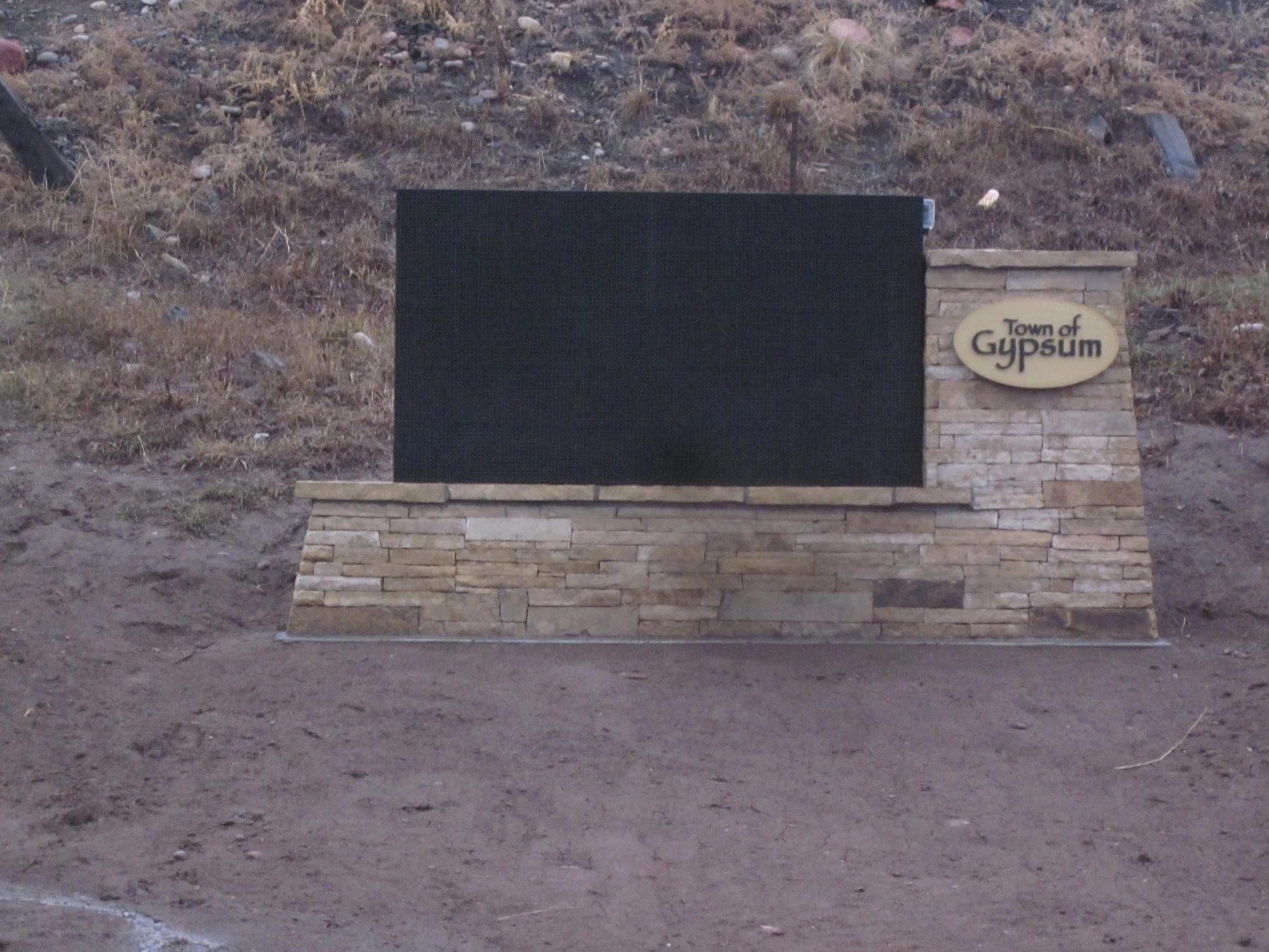 A sign on a stone structure that says 'Town of GypSum' with a large black blank board next to it, set against a dirt and hillside background.