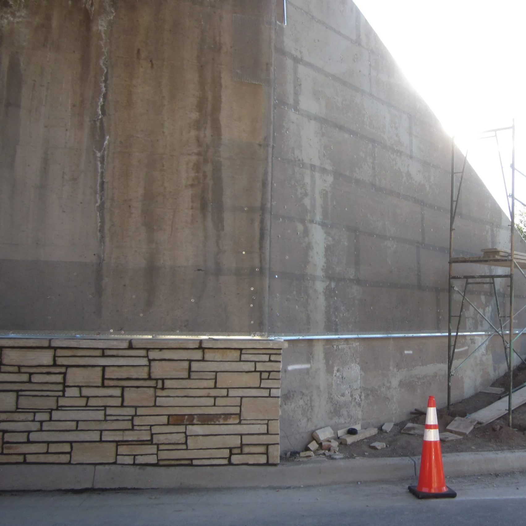 Side view of building construction with a concrete wall, metal scaffolding on the right, a stone brick wall detail at bottom left, construction cones, and debris on the ground.