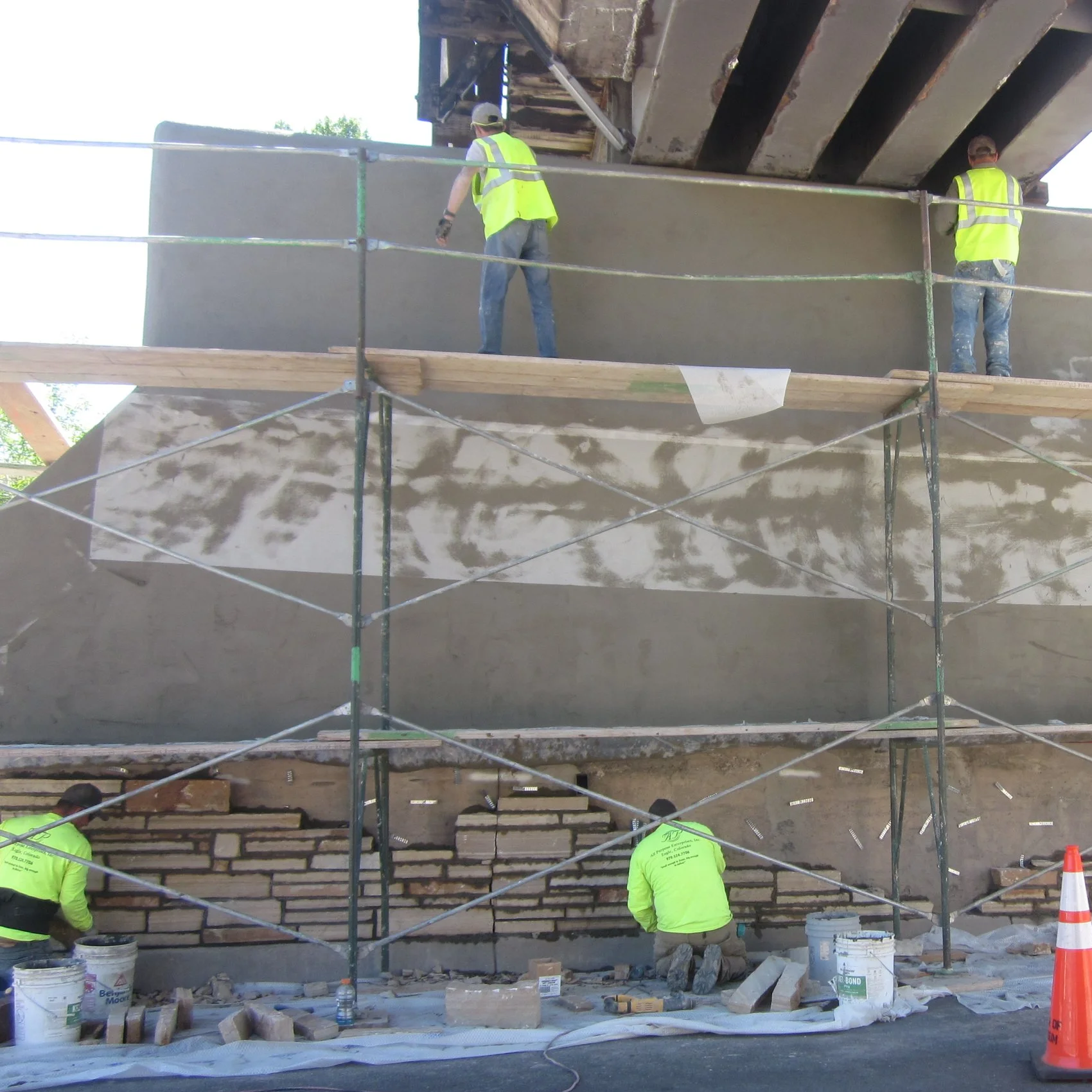 Construction workers wearing yellow safety vests and helmets working on a building with scaffolding, applying concrete or plaster on a wall.