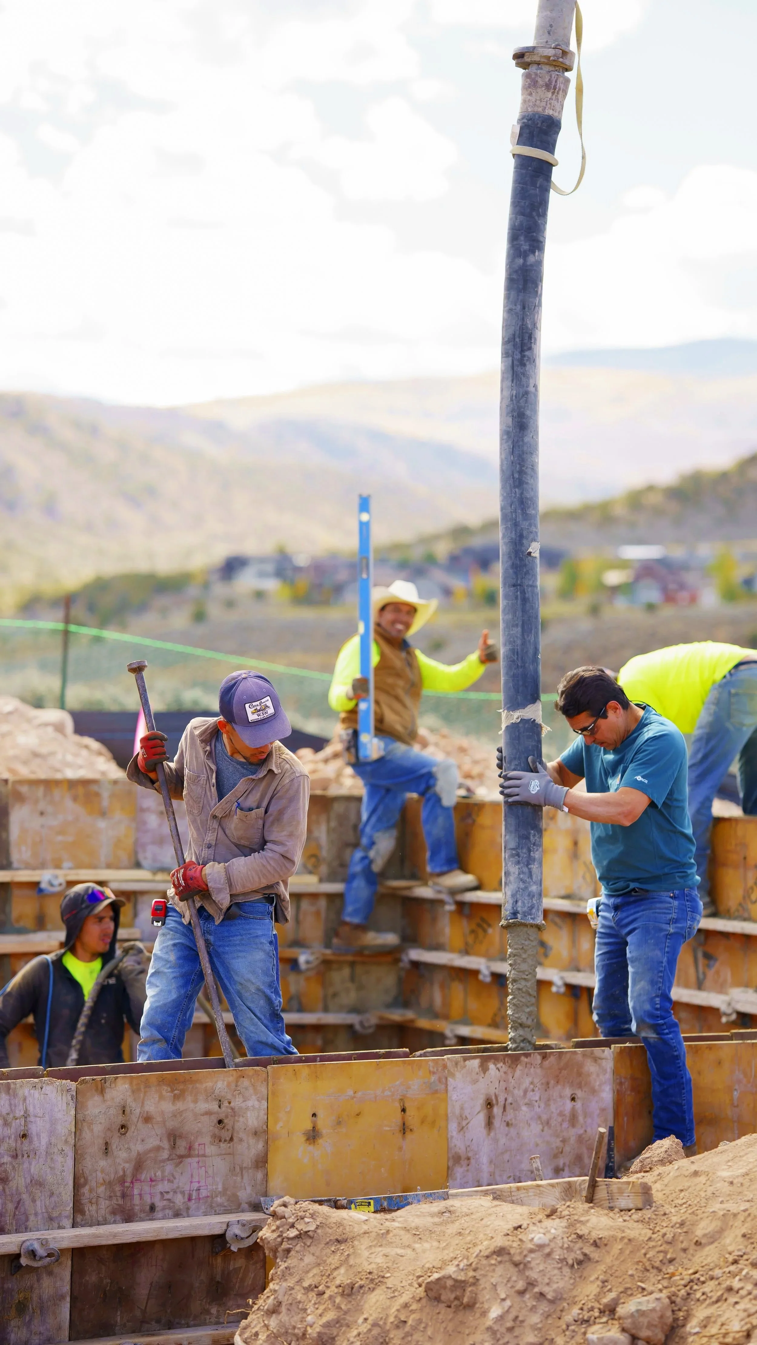 Construction workers pouring concrete and setting a steel pole at a construction site in a rural area with mountains in the background.