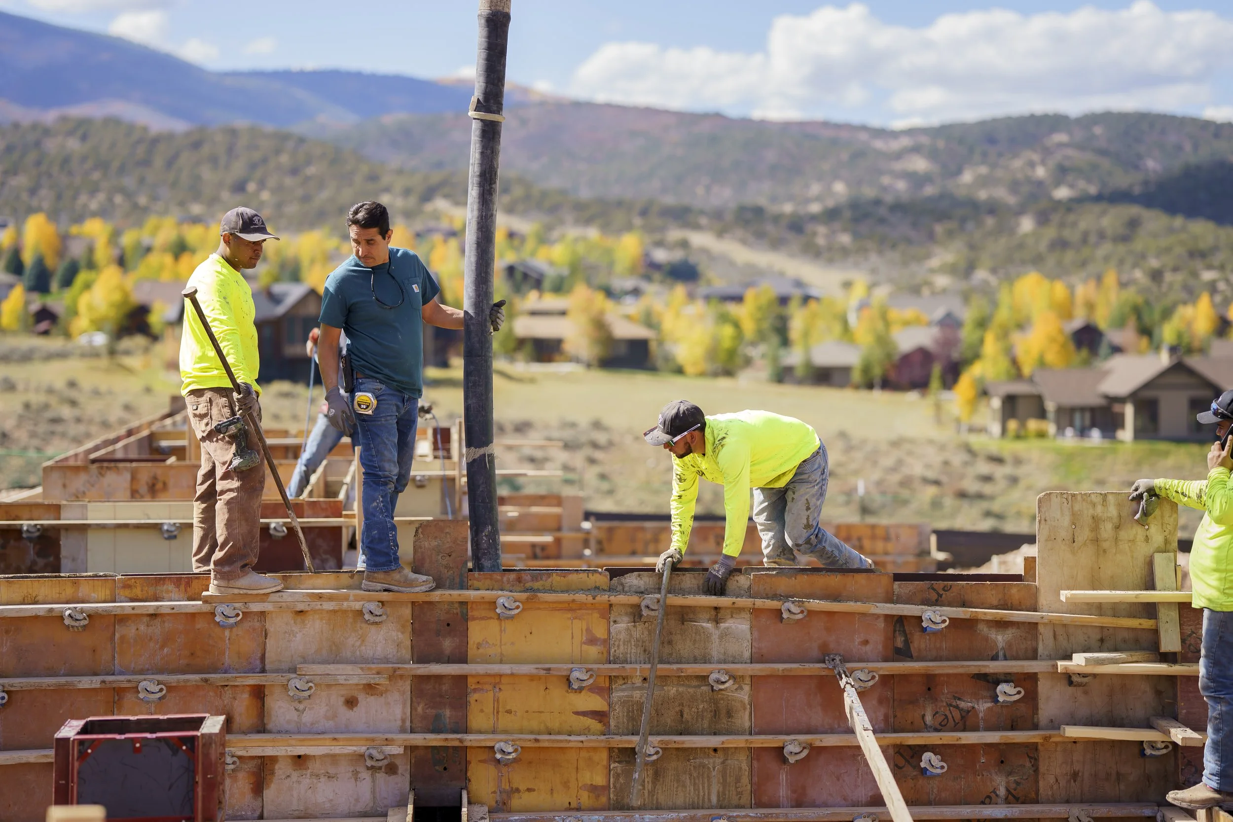 Construction workers pouring concrete into a foundation on a building site with a backdrop of mountains and trees with autumn foliage.