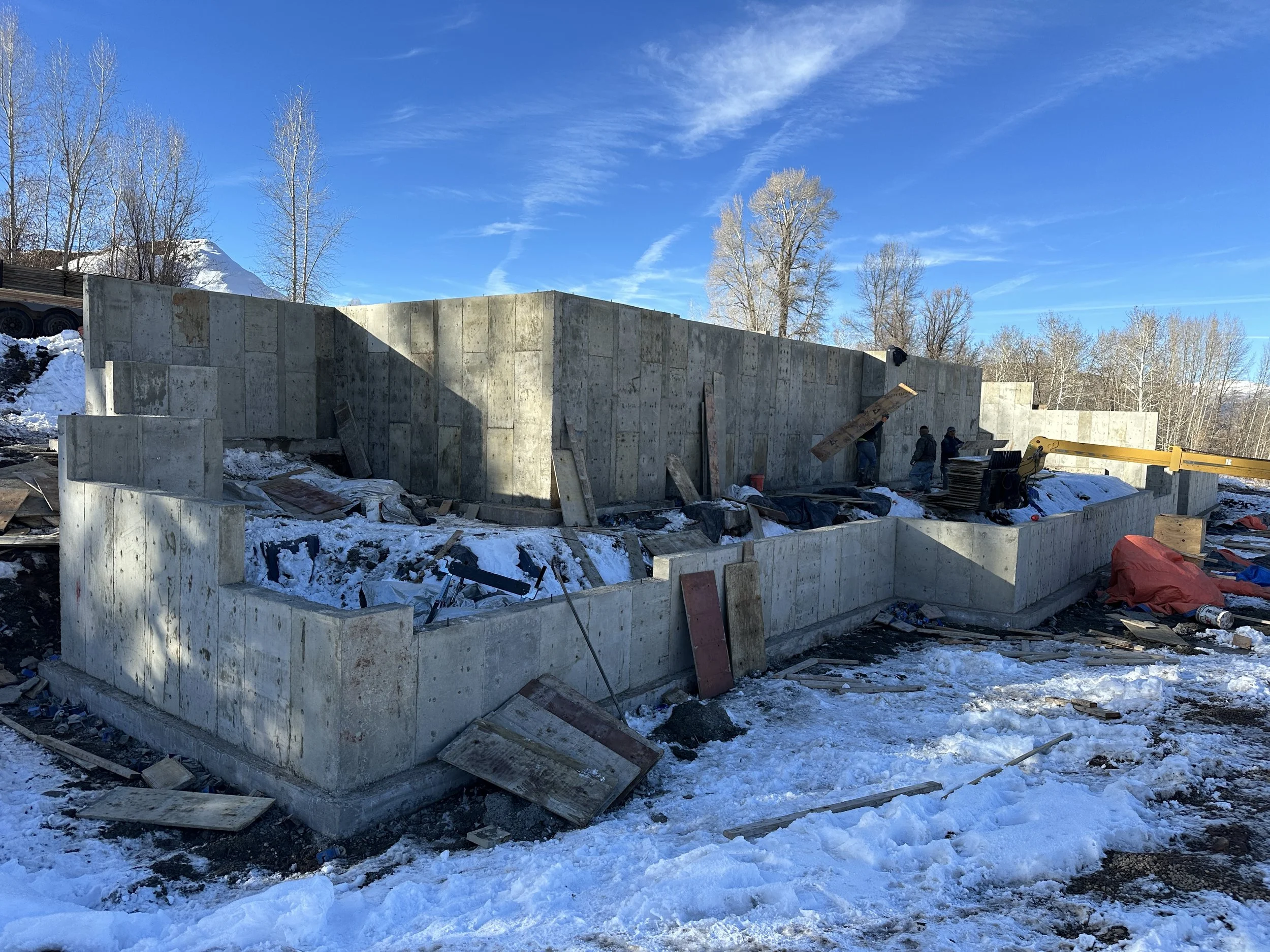 Construction site with concrete walls being built, surrounded by snow and with a clear blue sky. Workers and construction equipment are visible.