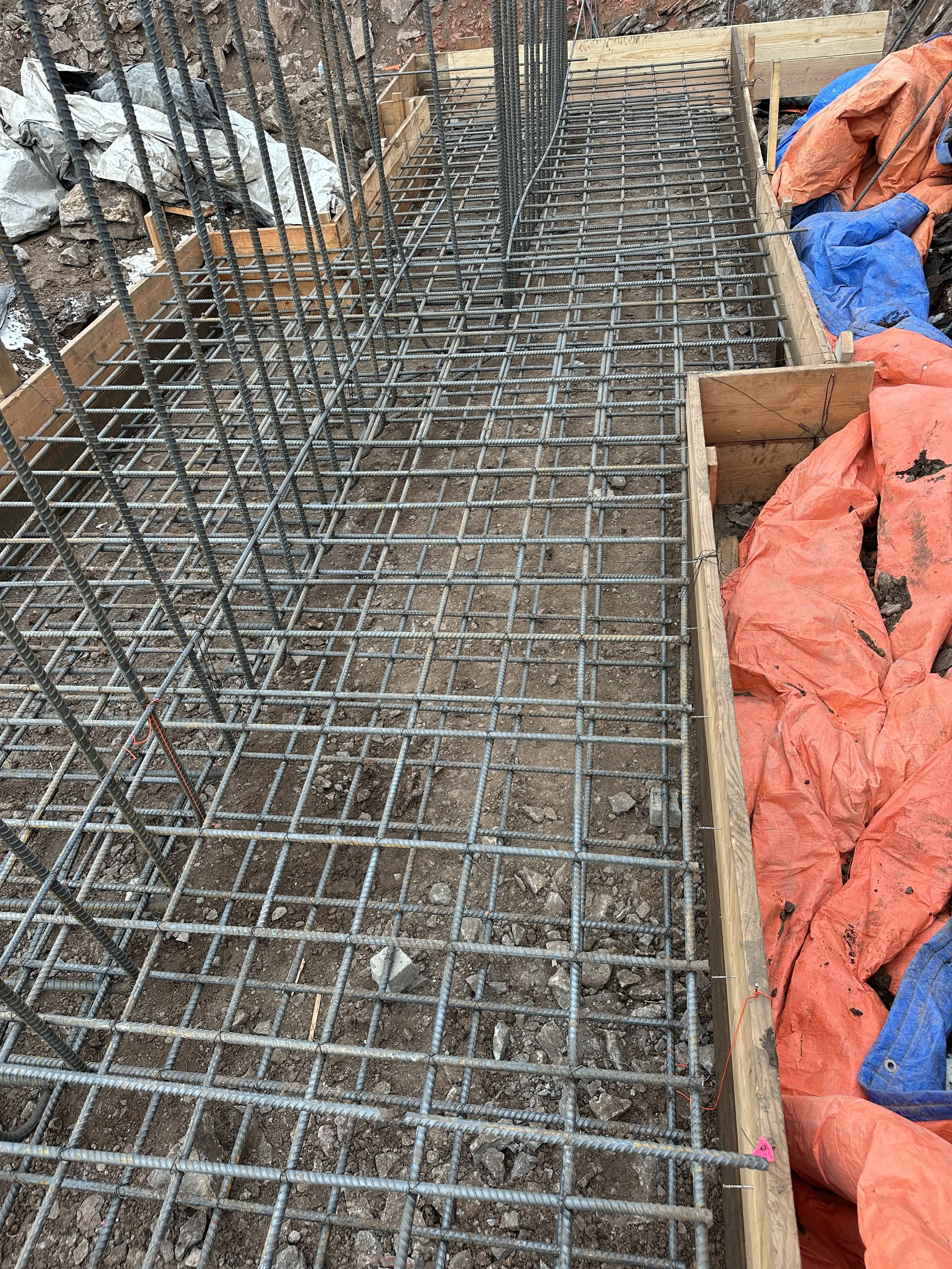 Construction site with steel rebar grid for concrete foundation, wooden formwork surrounding the area, and construction materials covered with orange and blue tarps.