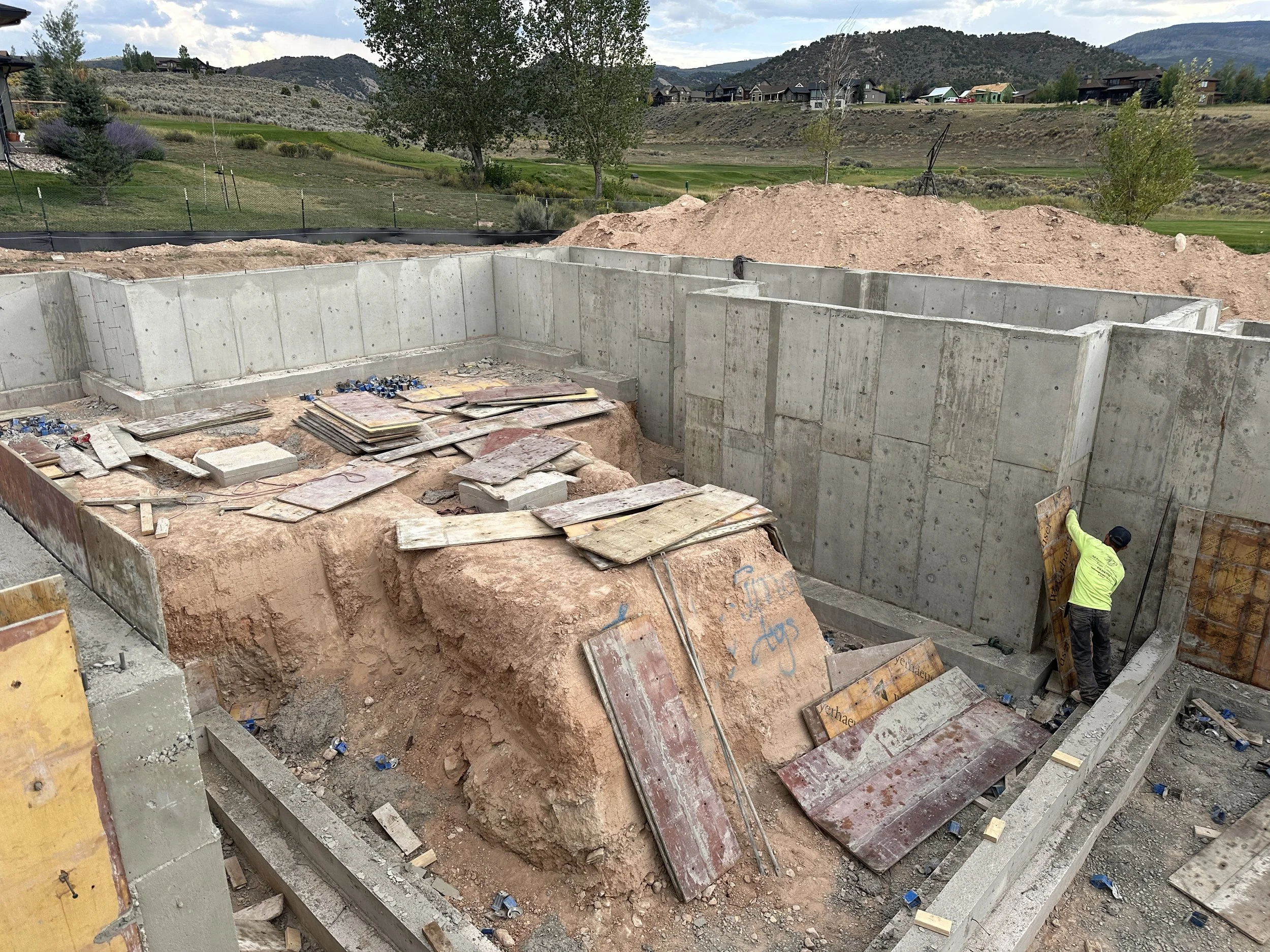 Construction site with concrete foundation walls and a worker installing form panels, with dirt and construction materials around, in a hilly area with trees and houses in the background.