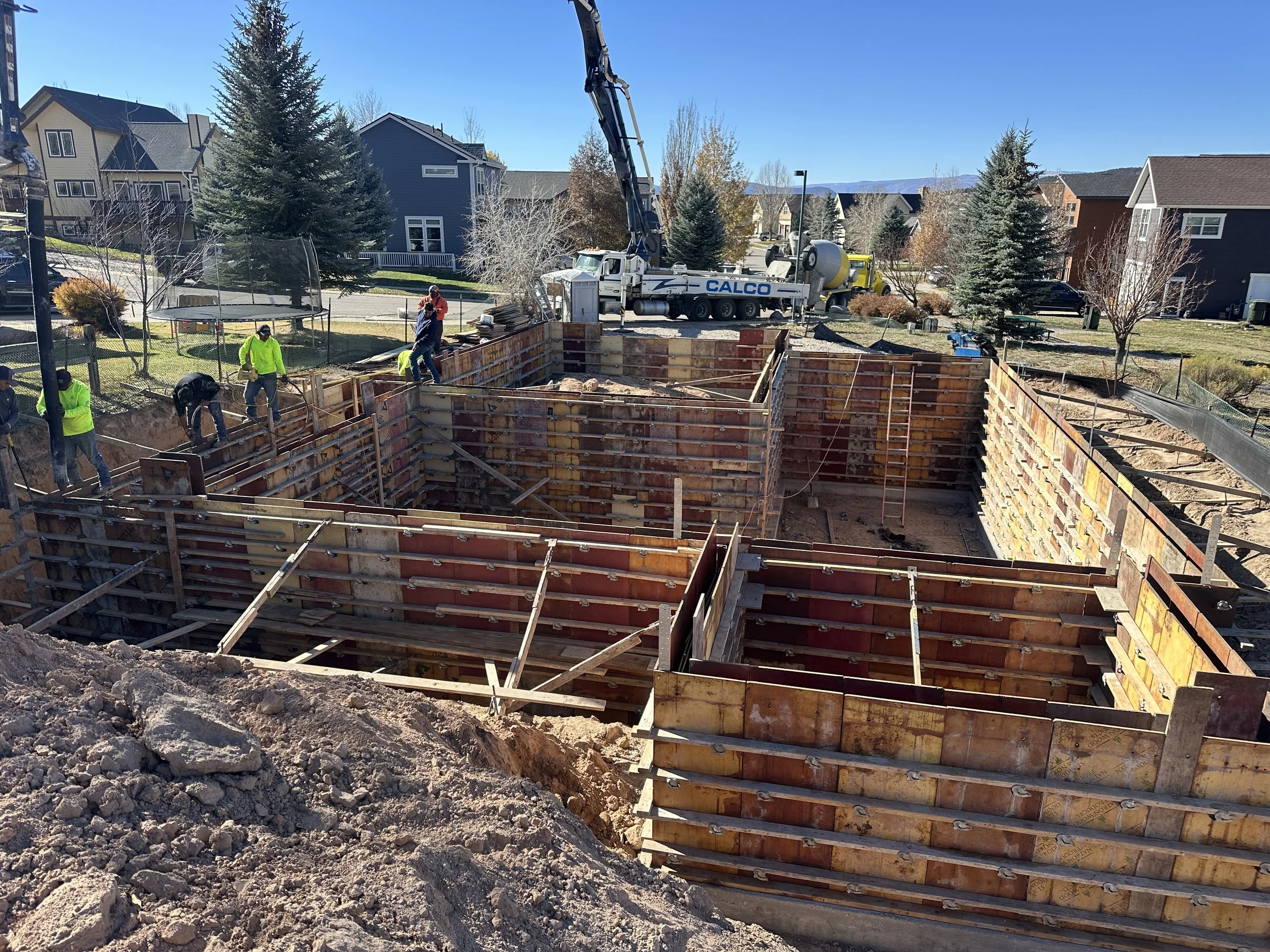 Construction workers at a building site with a concrete pump truck, working on the foundation of a structure in a residential neighborhood with houses and trees in the background.