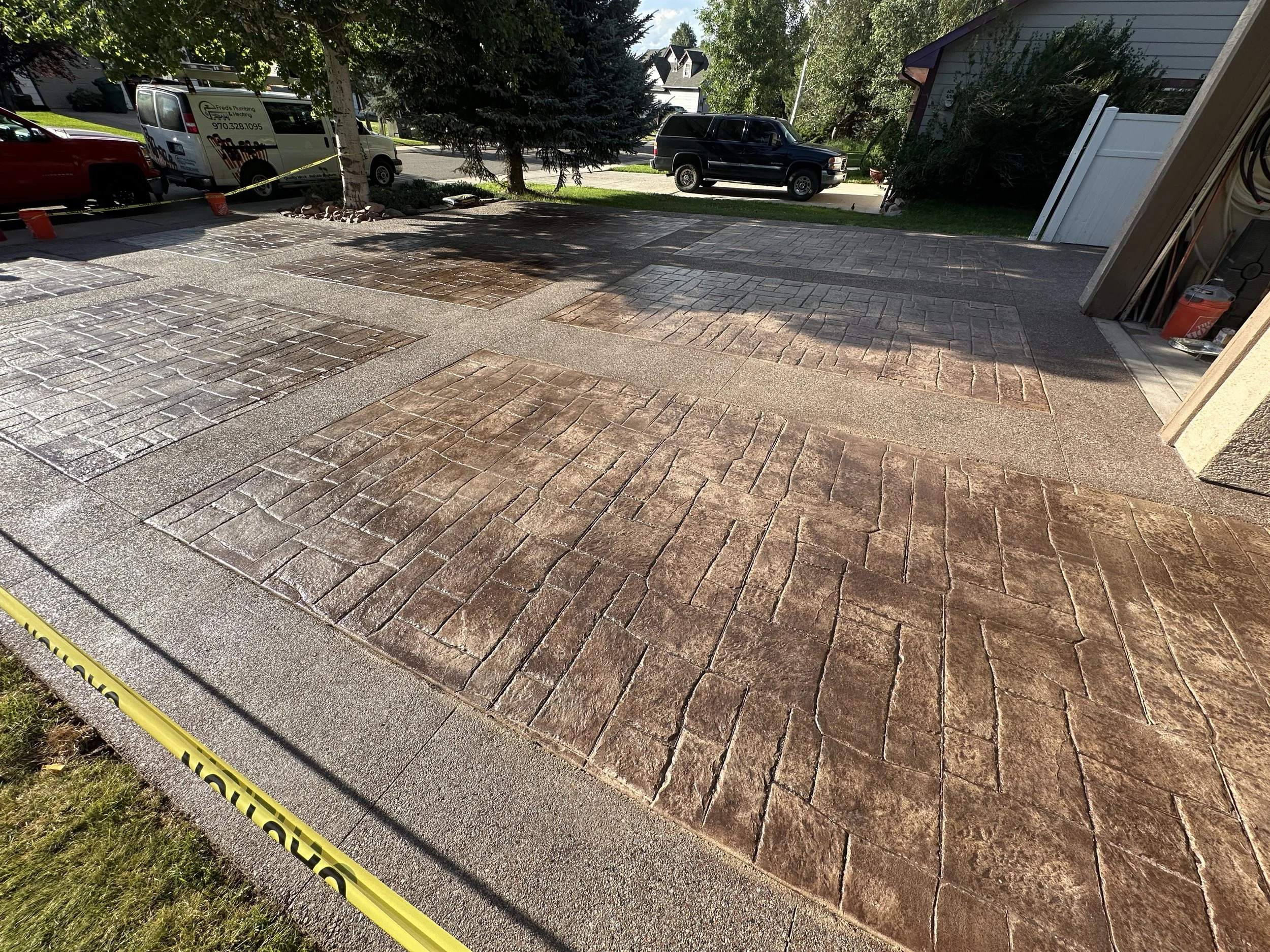 Newly paved driveway with decorative brick pattern, bordered by a yellow caution tape, with parked cars and trees in the background.