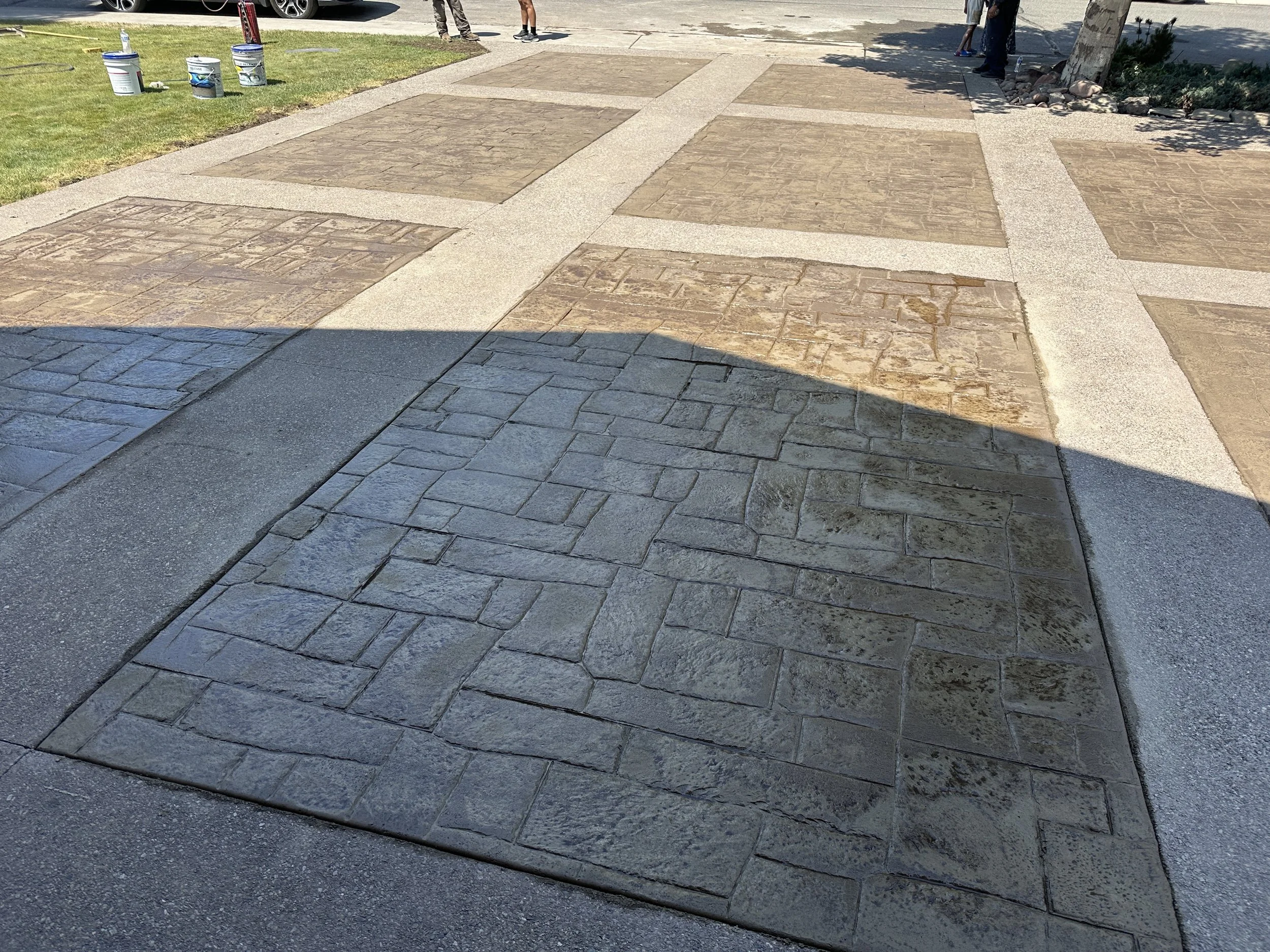A close-up view of a patterned concrete sidewalk with various textured paving stones, some with a brick-like appearance, and others with a smooth surface. A shadow partially covers the foreground of the sidewalk.