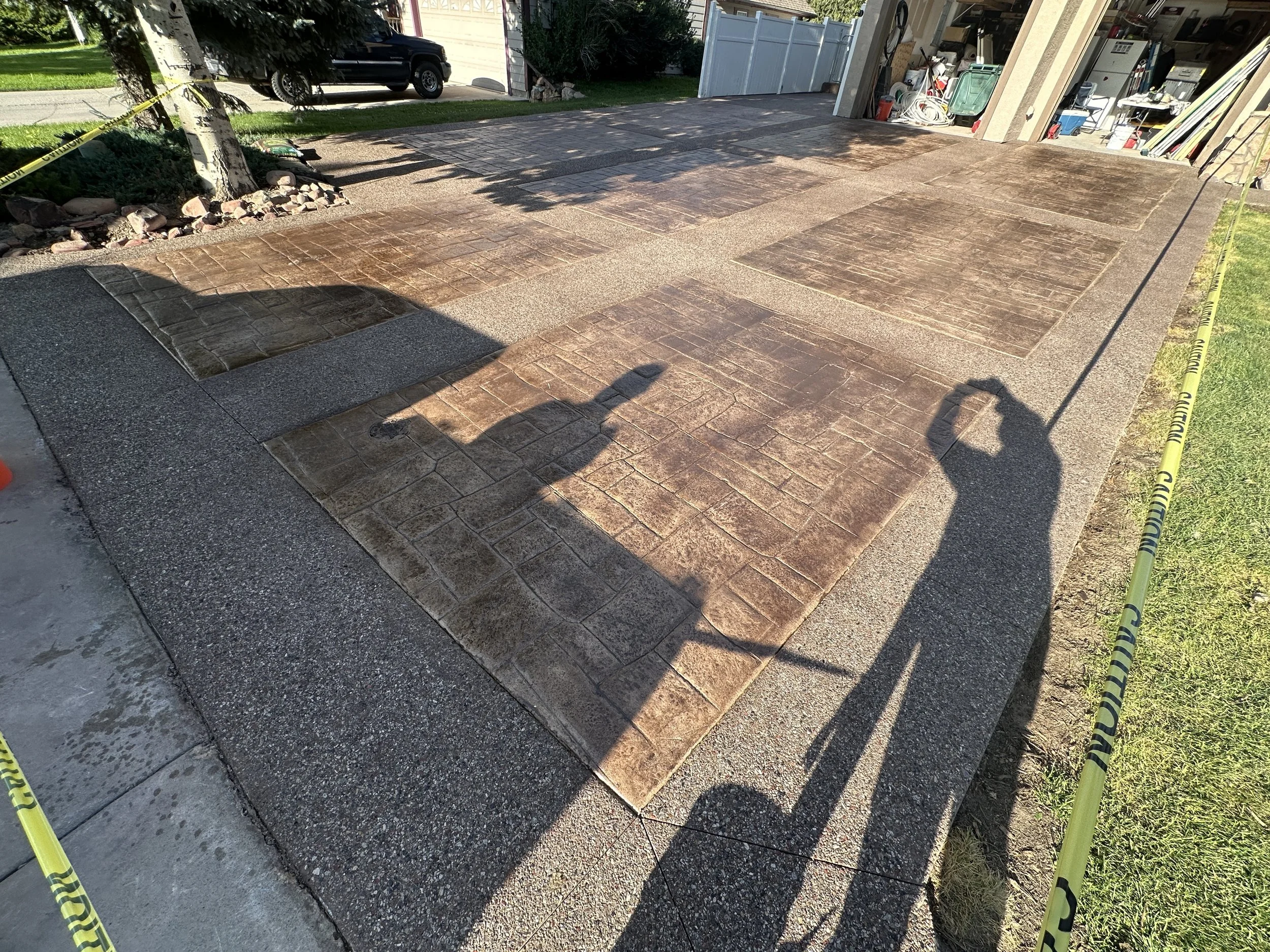 A newly paved walkway or driveway with brick and concrete patterns, surrounded by yellow caution tape, in front of a garage and house, with shadows of people and a street view in the background.
