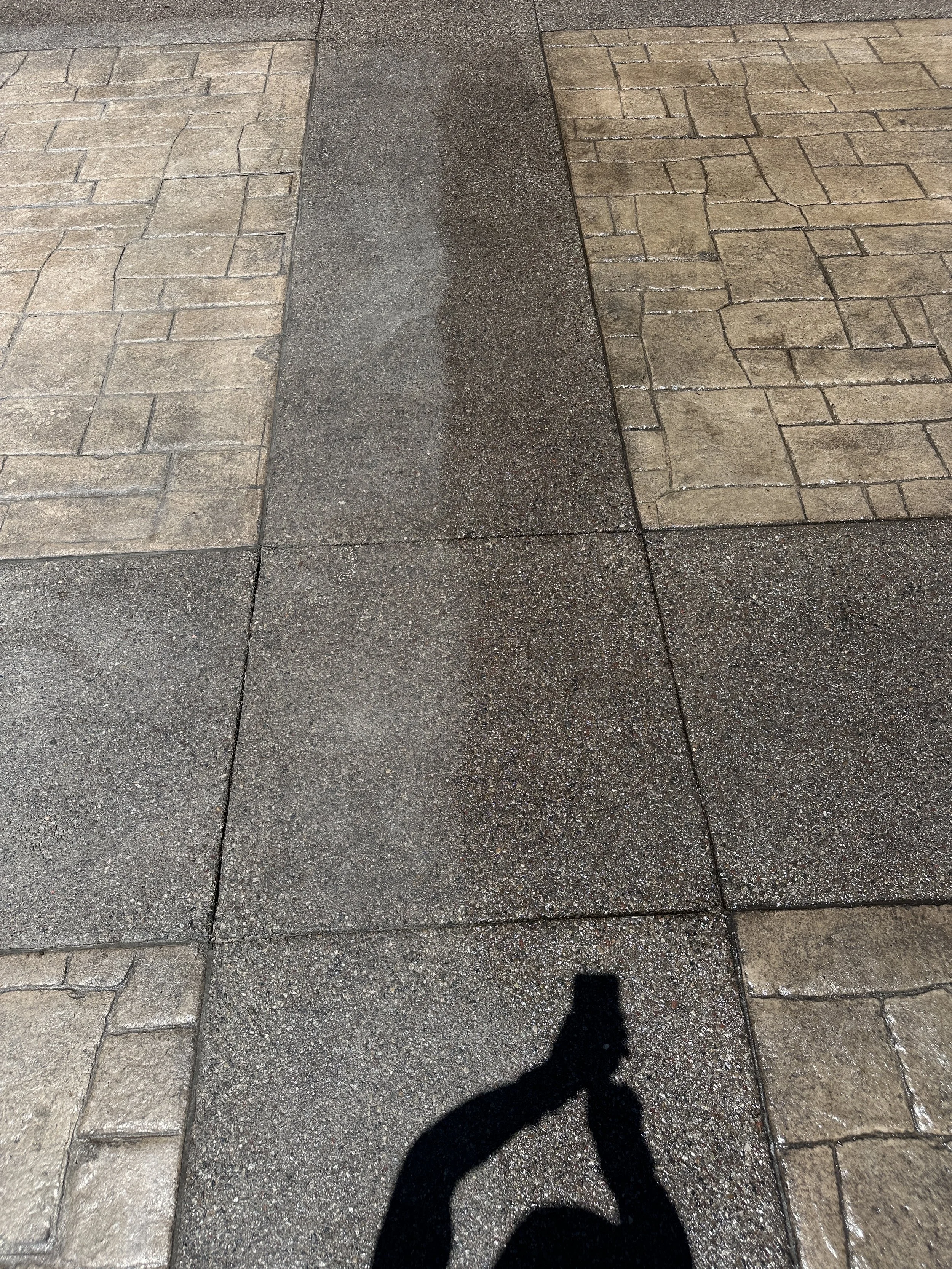Shadow of a person taking a photo, cast on a wet concrete sidewalk with different textured pavement sections.