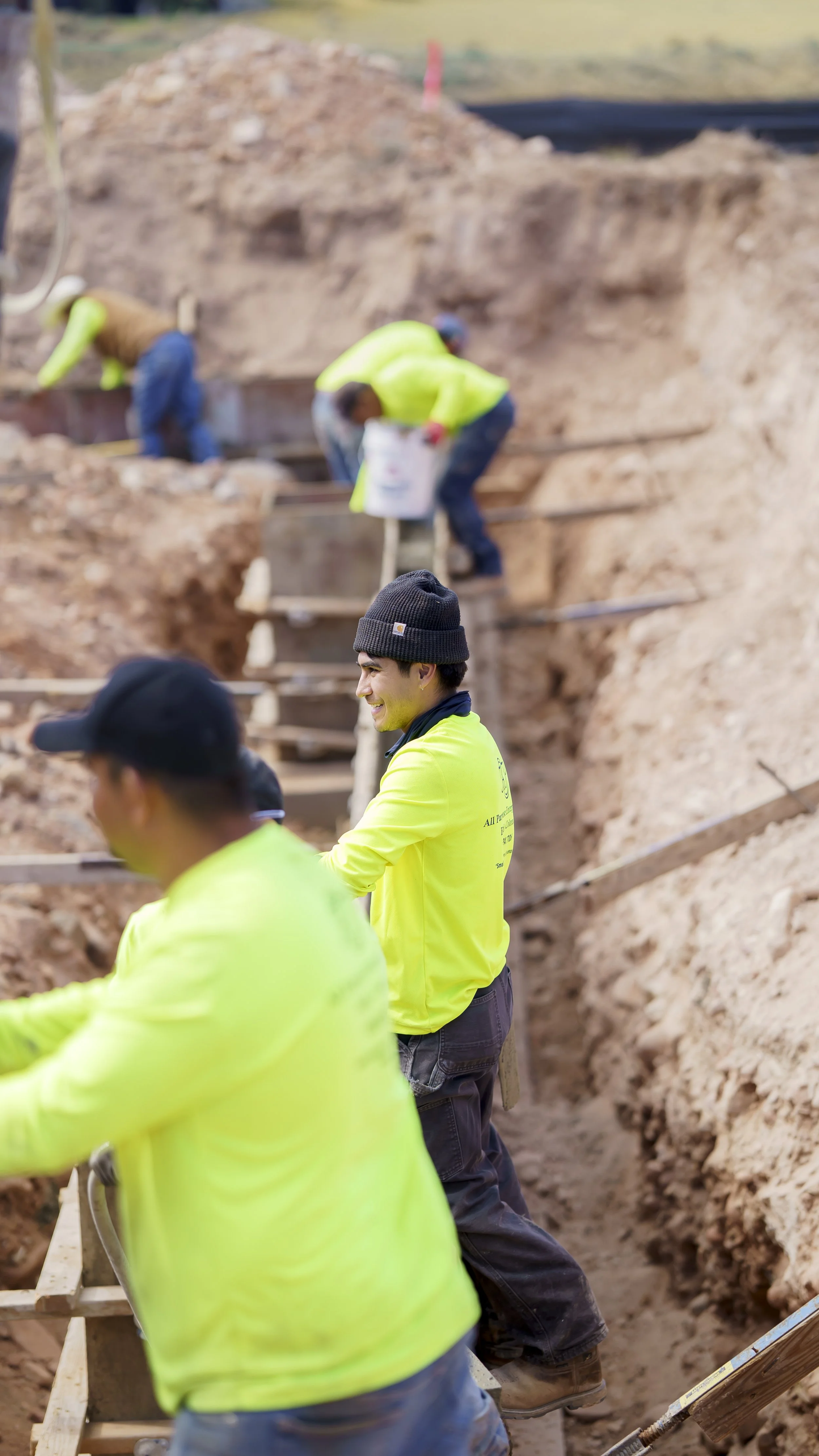 Construction workers in bright yellow shirts working on a site with dirt and wooden scaffolding.