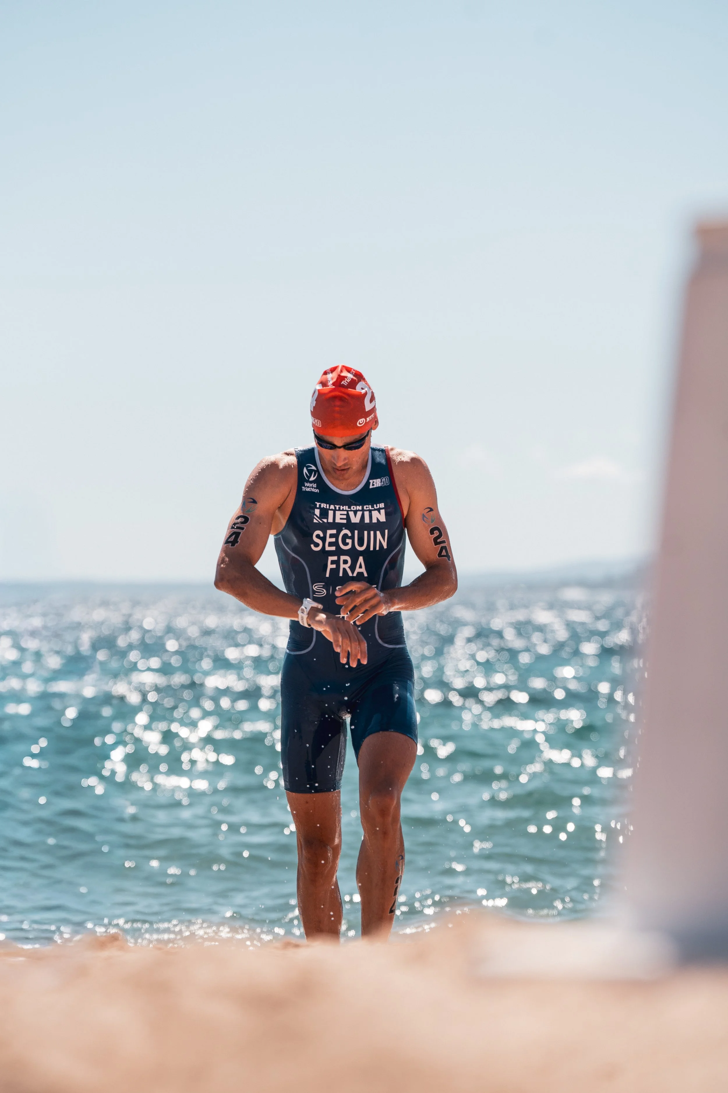 Triathlète homme portant un maillot de triathlon noir avec texte blanc, casque rouge, lunettes de soleil, courant sur la plage avec la mer en arrière-plan, sous un ciel clair et ensoleillé.