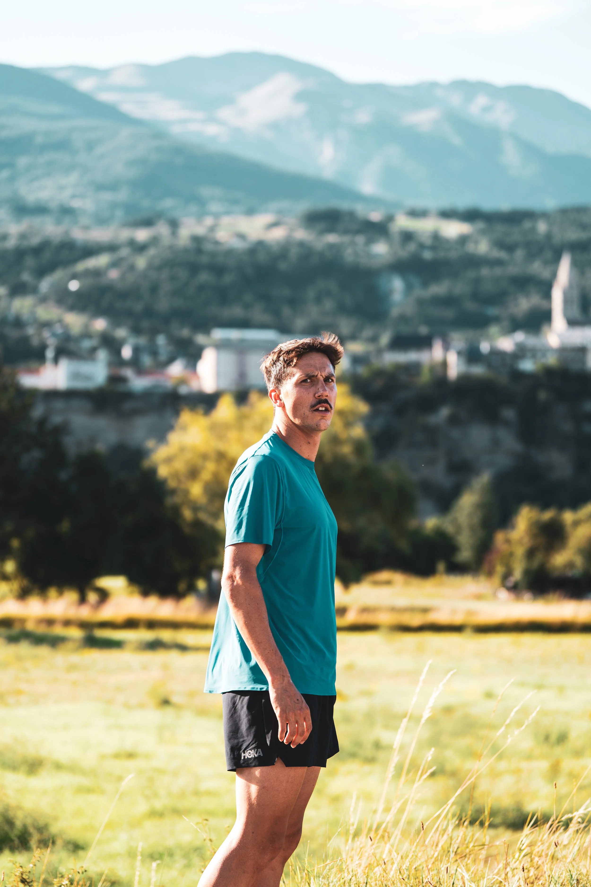 Un homme en t-shirt bleu et short noir se tient dans un champ en plein air avec des montagnes en arrière-plan.