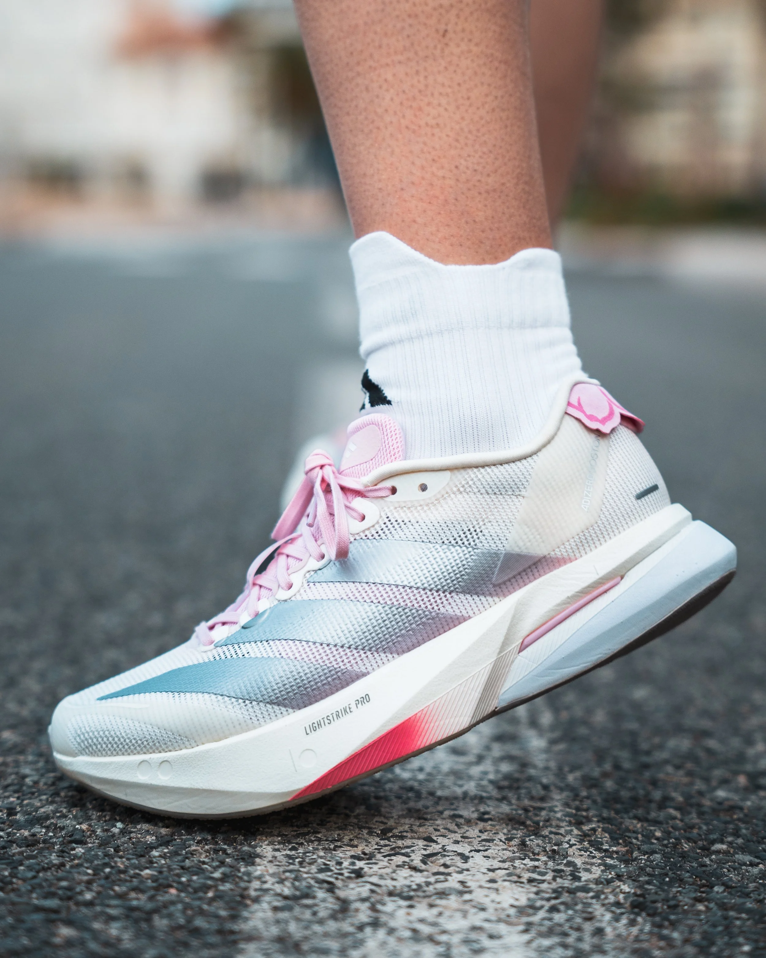 Close-up d'une chaussure de course blanche et rose avec une semelle épaisse, portée par une personne dont la jambe est visible, vêtue d'une chaussette blanche. La chaussure est utilisée sur une surface asphaltée.