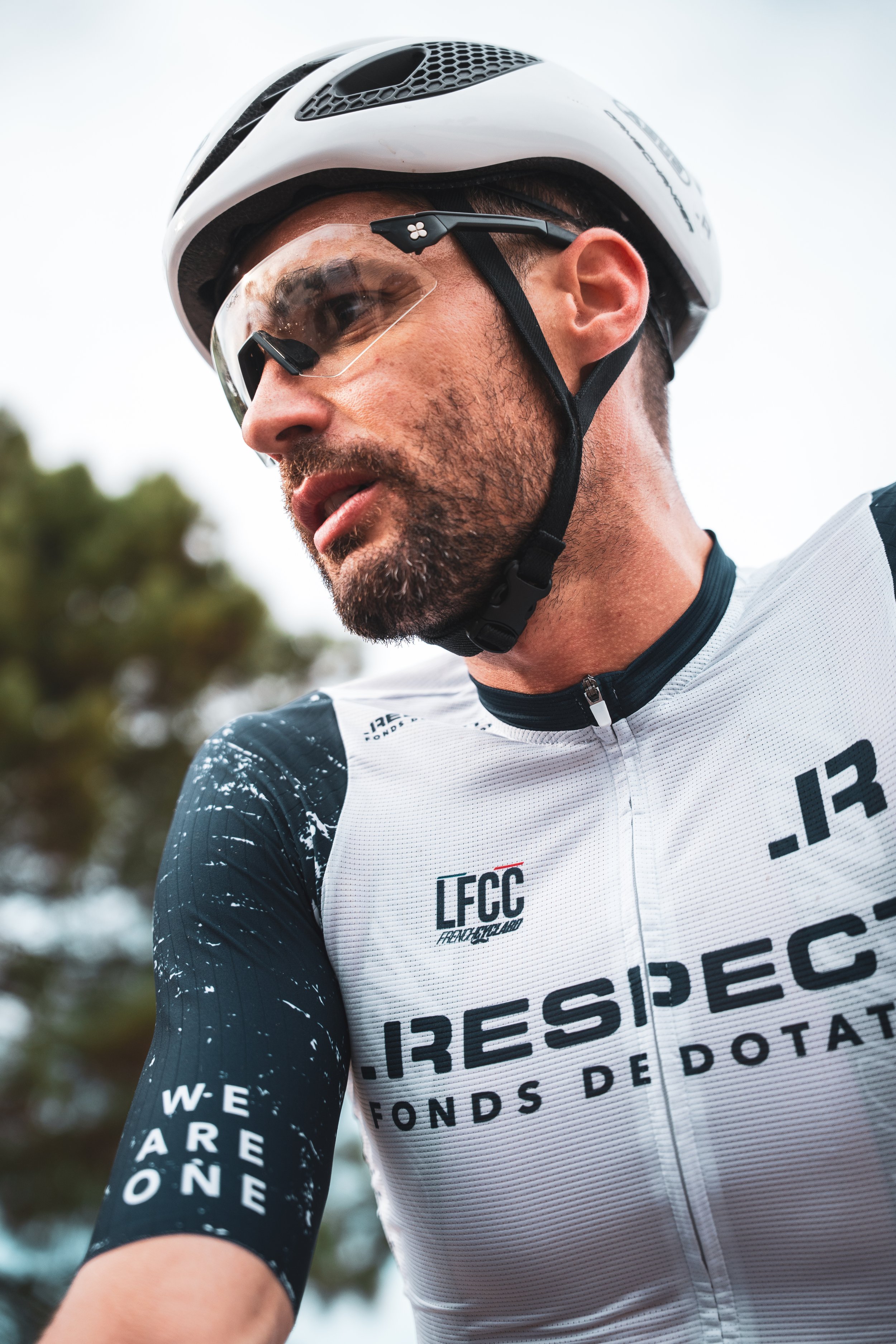 Un homme portant un casque de vélo, des lunettes de protection et une tenue de cyclisme, en plein effort lors d'une sortie en extérieur, avec des arbres en arrière-plan.