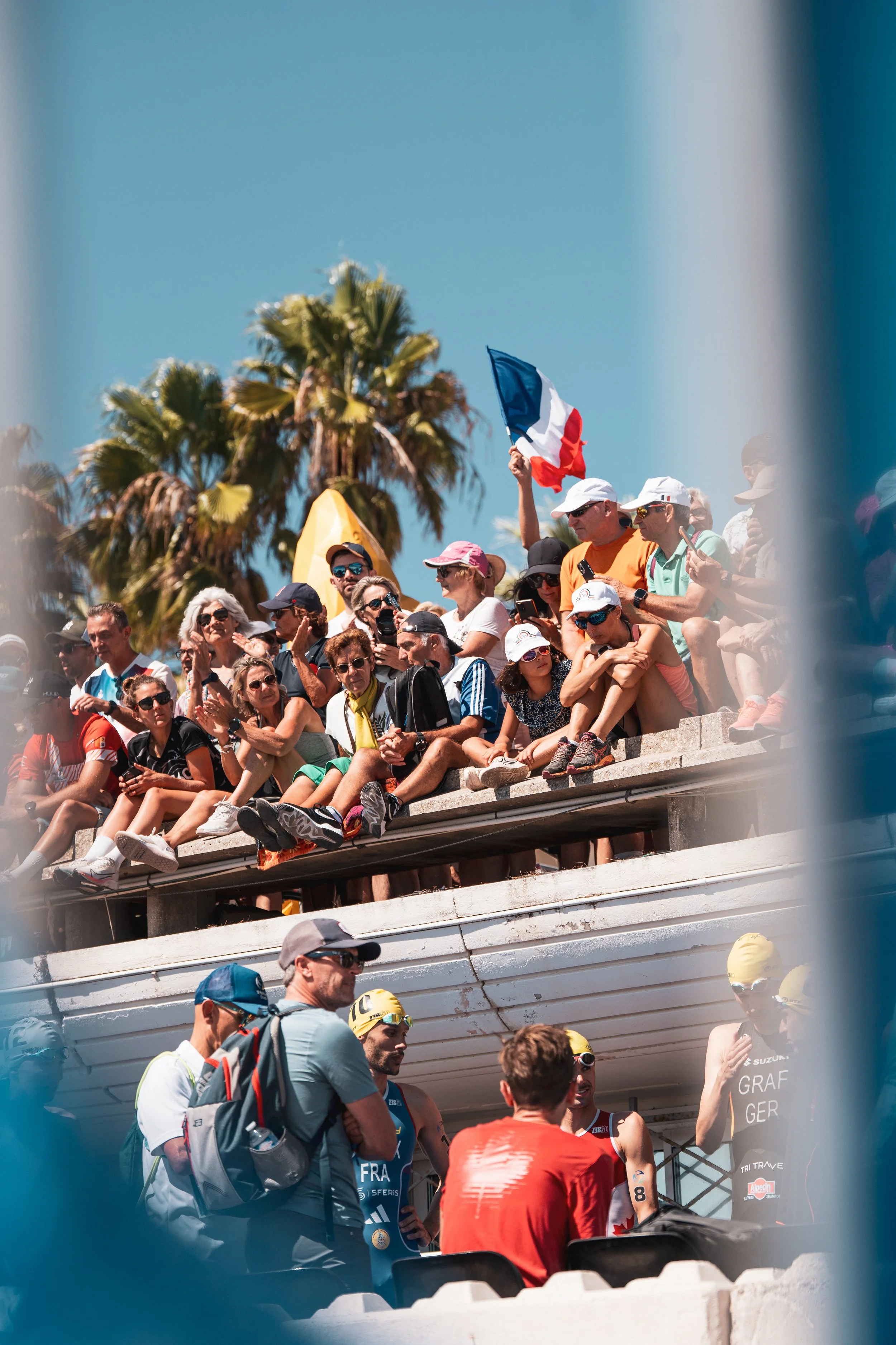 Groupe de personnes lors d'un événement sportif en plein air, avec un drapeau français et des palmiers en arrière-plan.