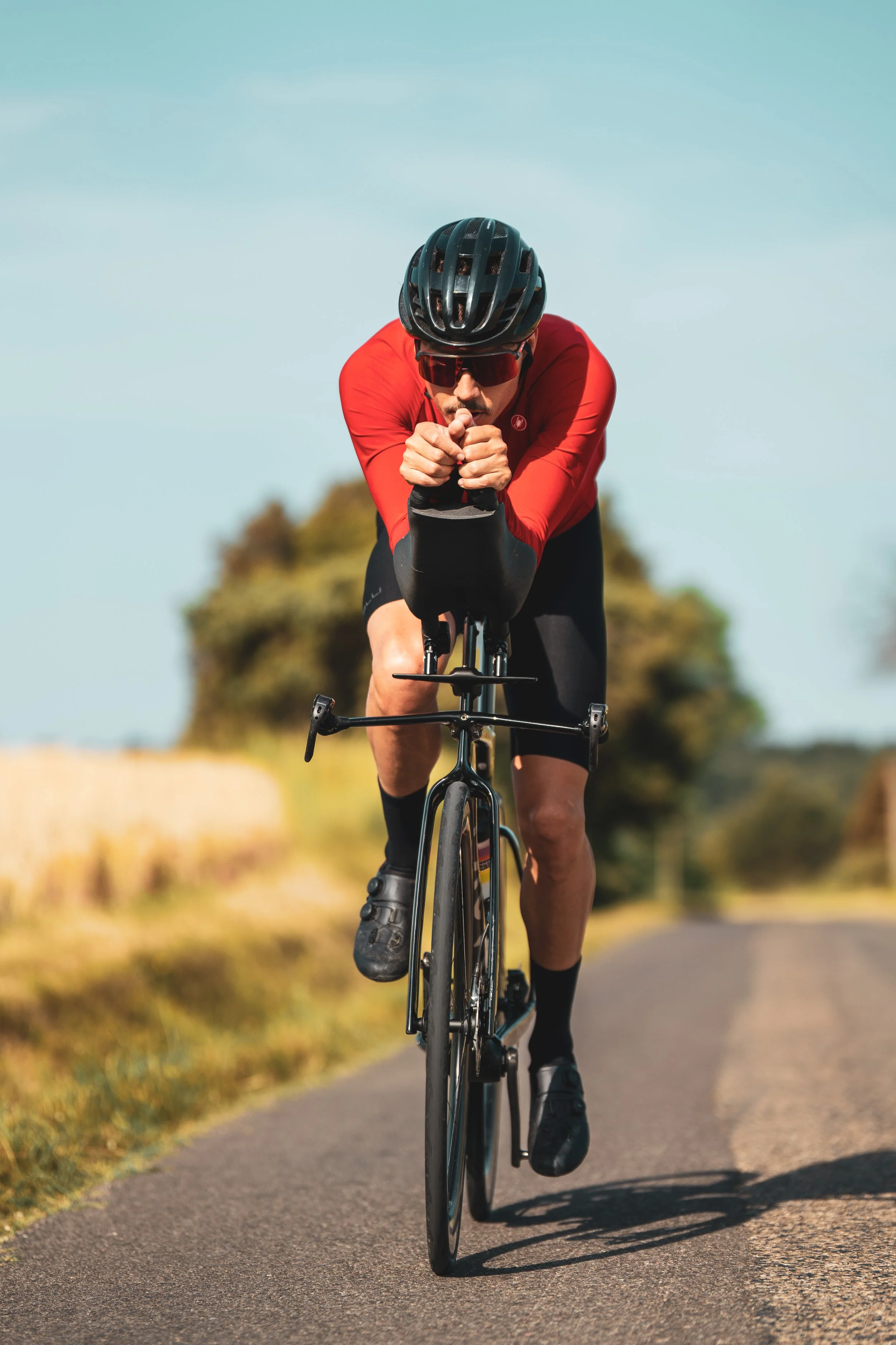Un cycliste portant un casque noir, des lunettes de soleil rouges et une veste rouge, pédale sur une route pavée en plein air par une journée ensoleillée.