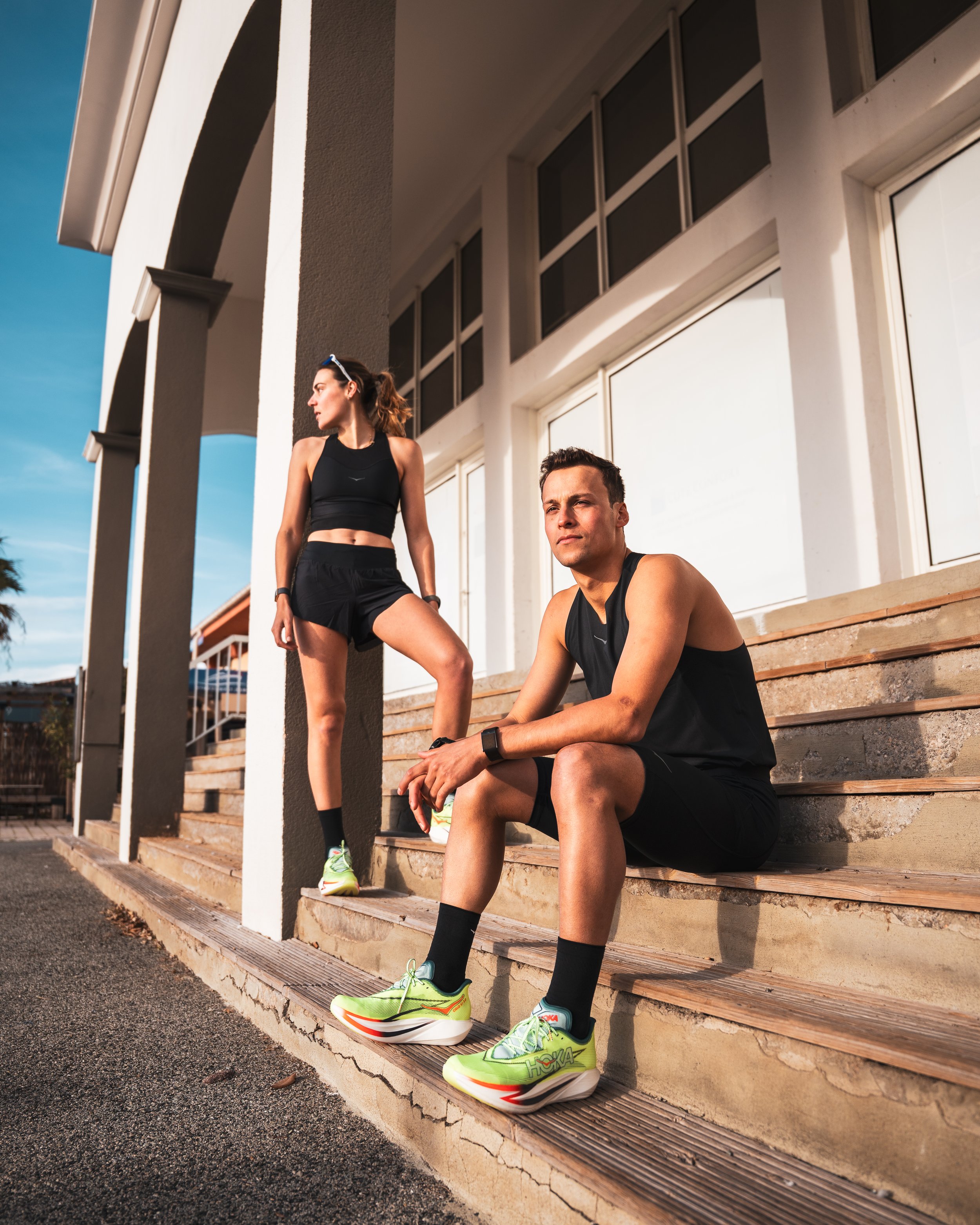 Deux personnes en tenue de sport s'entraînent en extérieur sur un escalier en bois, avec un bâtiment moderne en arrière-plan, lors d'une séance de fitness ou de course.