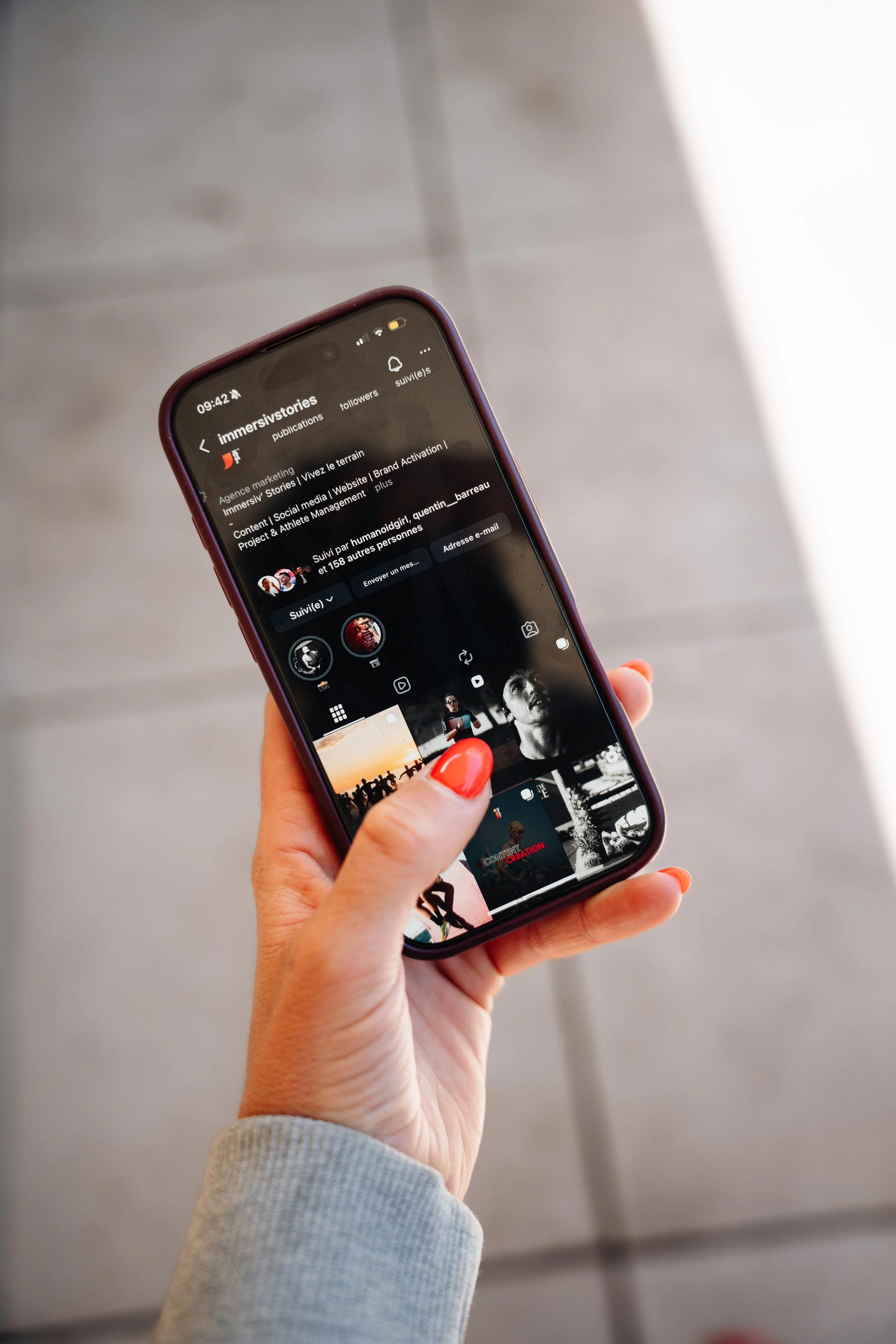 Une main tenant un smartphone affichant une page de profil sur une application de réseaux sociaux, avec des images en bas de l'écran et un fond flou.
