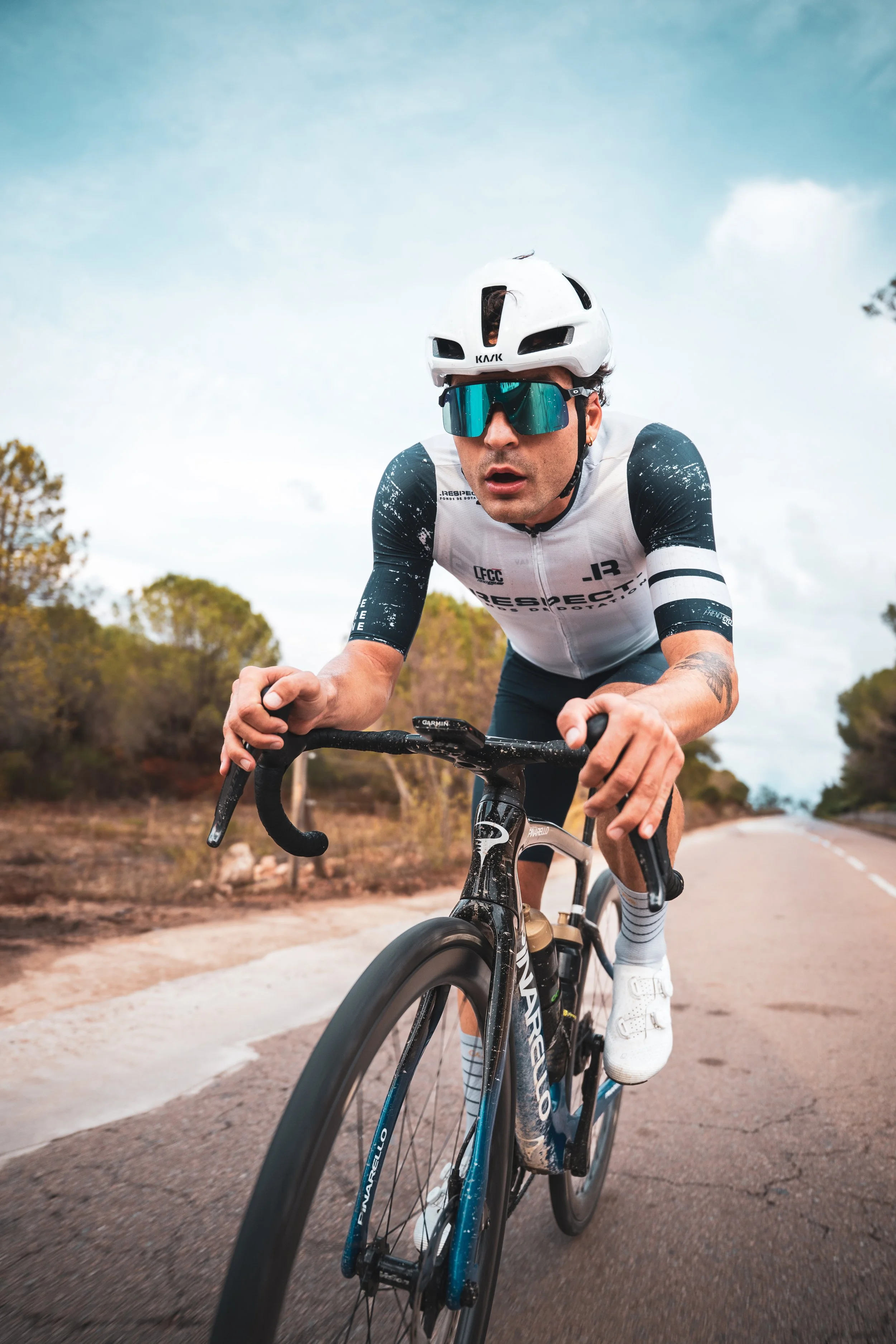 Cycliste en tenue de sport portant un casque blanc et des lunettes de soleil, pédalant sur une route pavée avec des arbres en arrière-plan, sous un ciel partiellement nuageux.