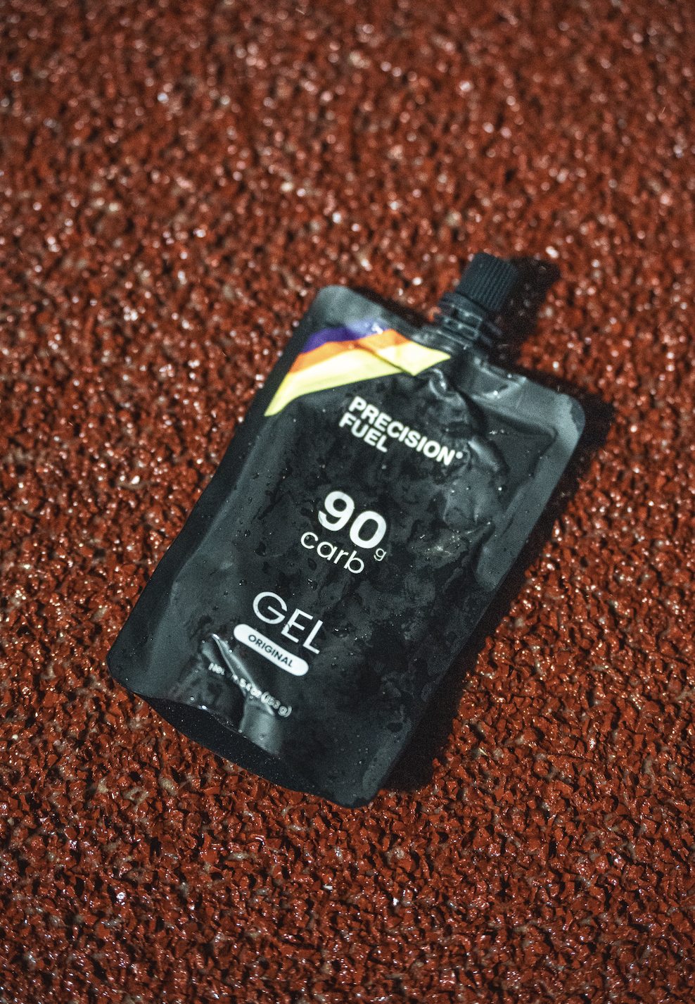 Sachet noir de gel de carburant Precision Fuel contenant 90 g de carb, posé sur une surface rouge granuleuse.
