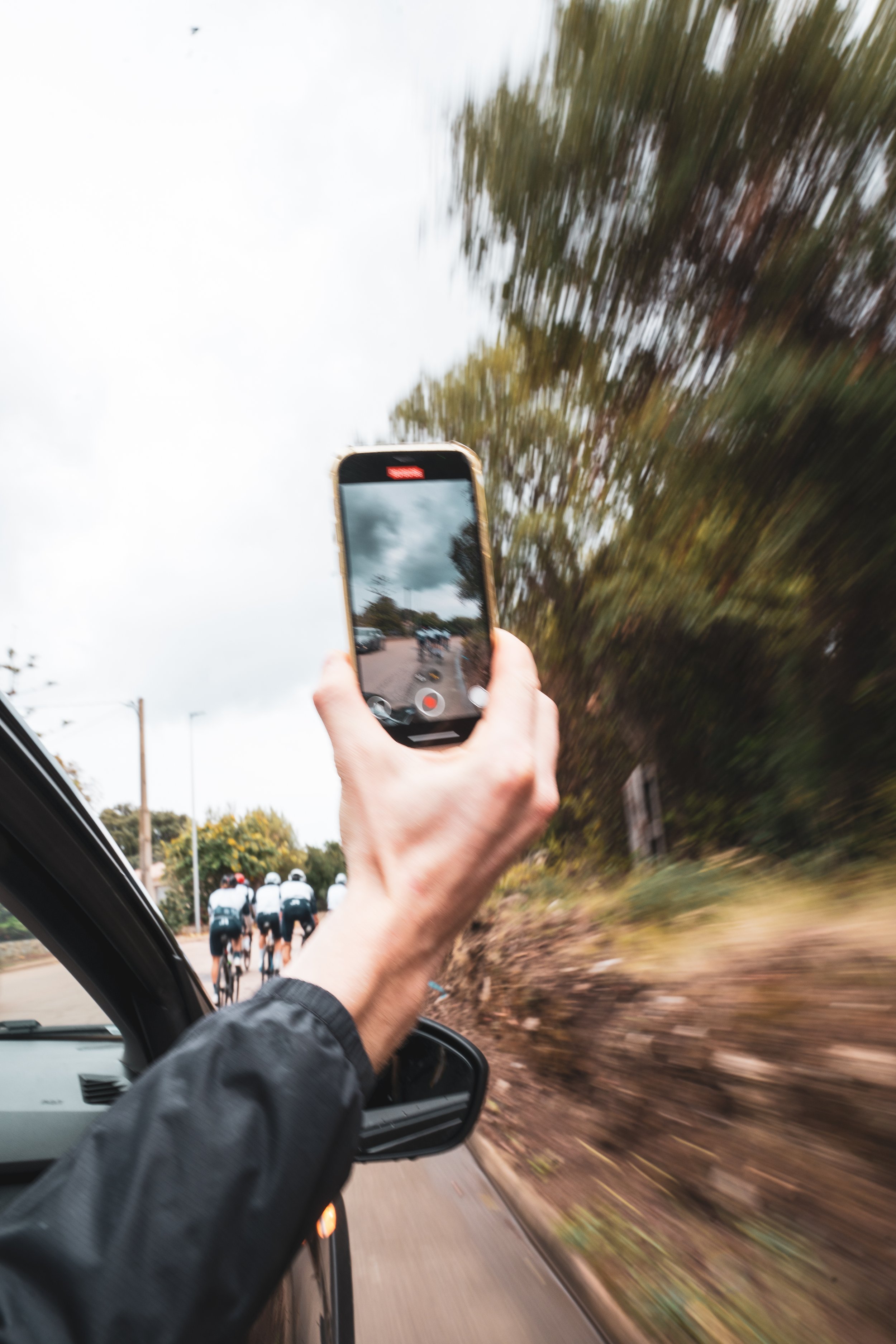 Une personne filme un groupe de cyclistes avec un téléphone depuis une voiture en mouvement, avec un arrière-plan de ciel nuageux et de végétation.