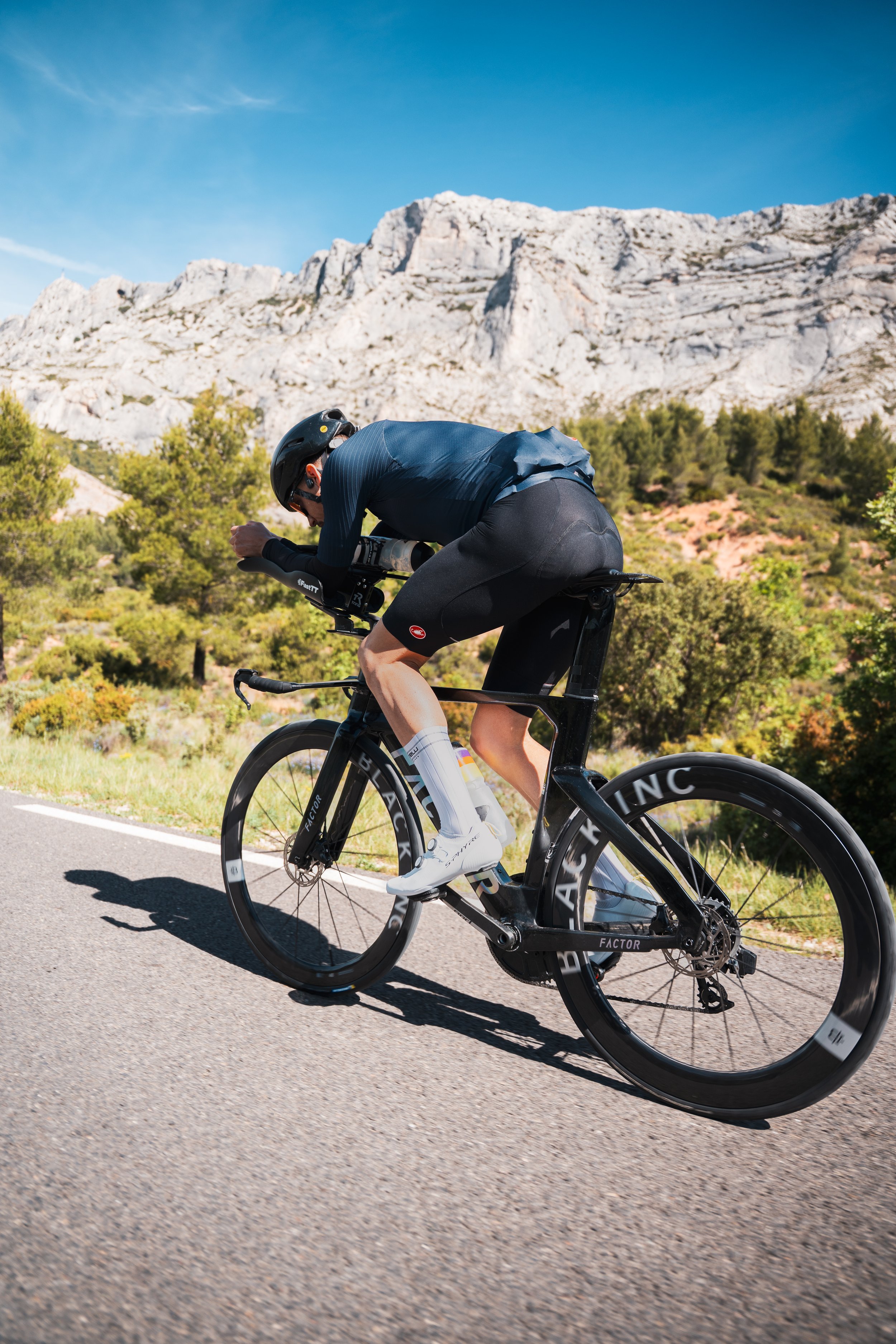 Un cycliste en tenue de course pédale sur une route, avec des montagnes rocheuses en arrière-plan et un ciel bleu clair.