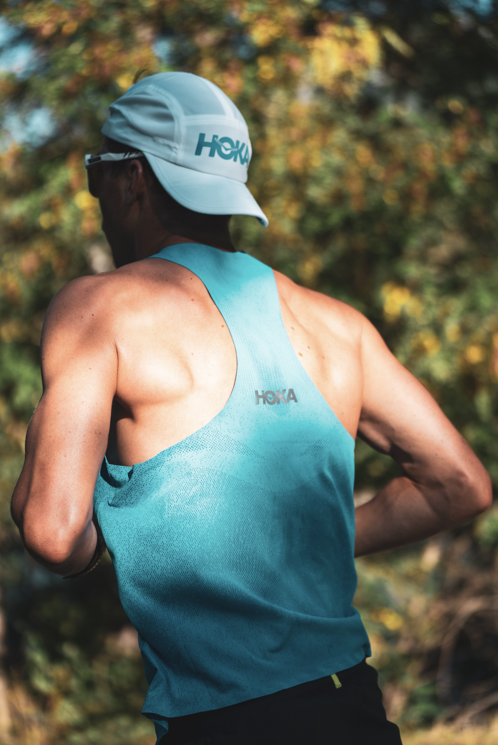 Un homme participe à une course ou séance de course à pied en portant une casquette blanche et un débardeur bleu turquoise avec le logo HOKA, dans un environnement naturel avec des arbres aux feuilles d'automne.