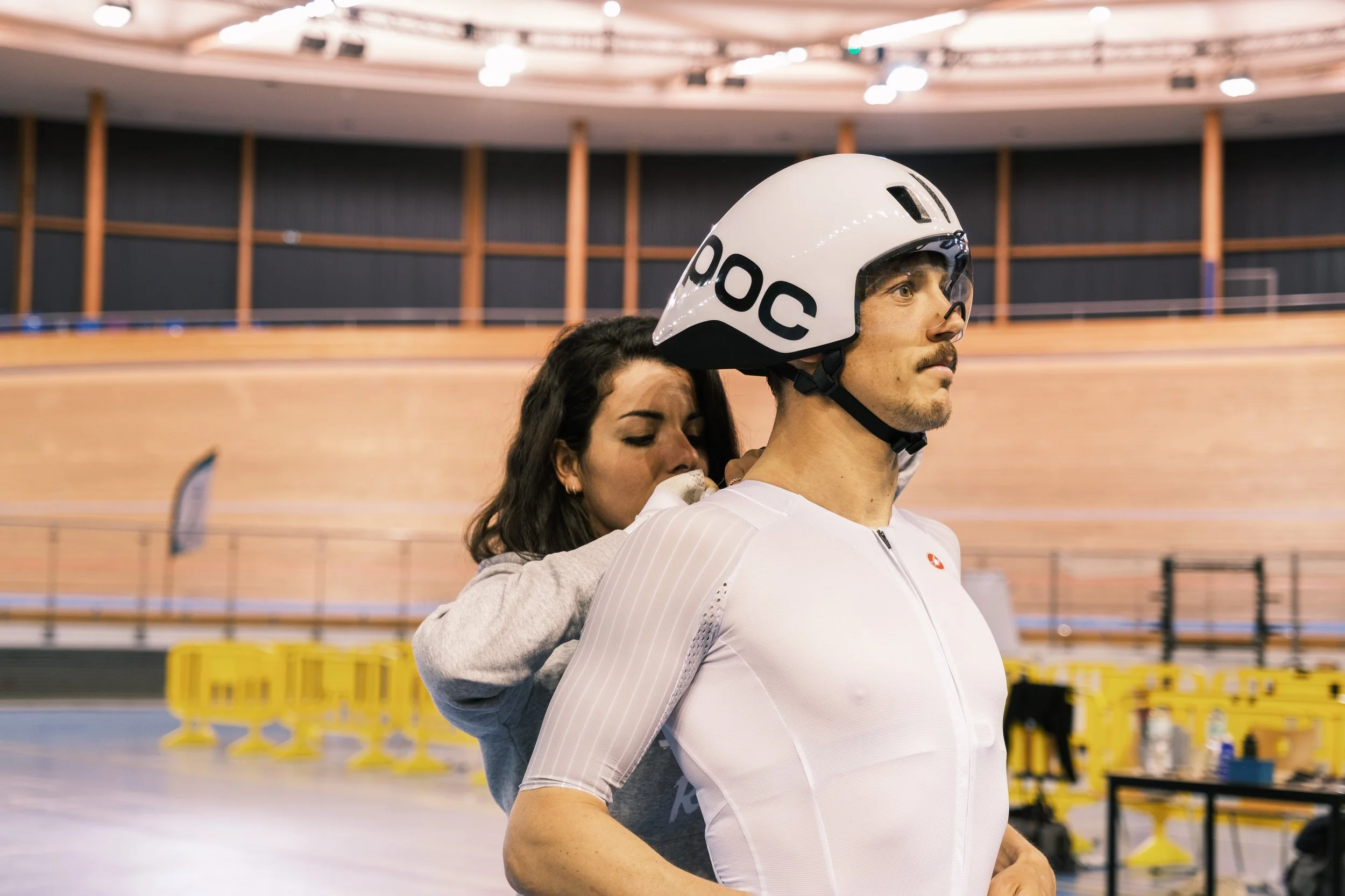 Un homme portant un casque de protection blanc avec le logo POOC, assis sur le dos d'une femme lors d'une séance dans un espace intérieur.