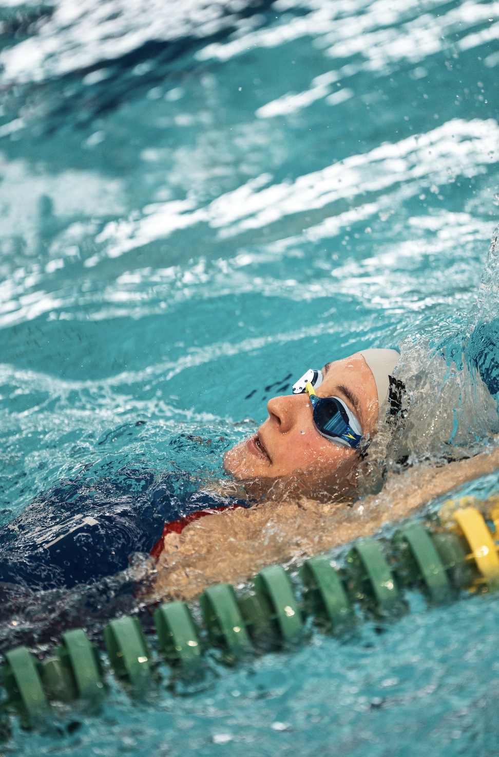 Un nageur en maillot rouge nageant en piscine, équipé de lunettes de natation et d'une bonnet blanc, près d'une ligne de départ verte.