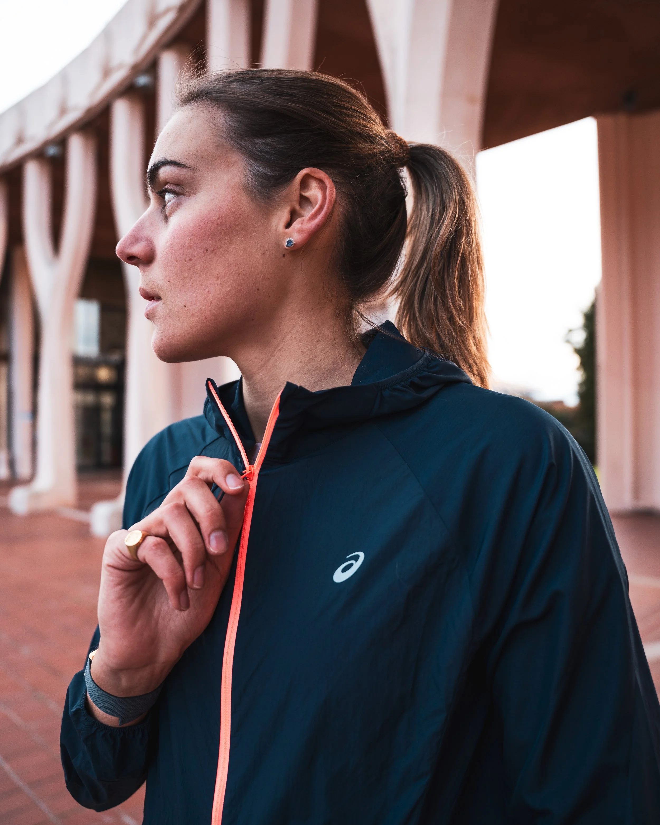 Femme portant un vêtement de sport, serrant une fermeture éclair orange sur une veste noire, avec une architecture en pierre en arrière-plan