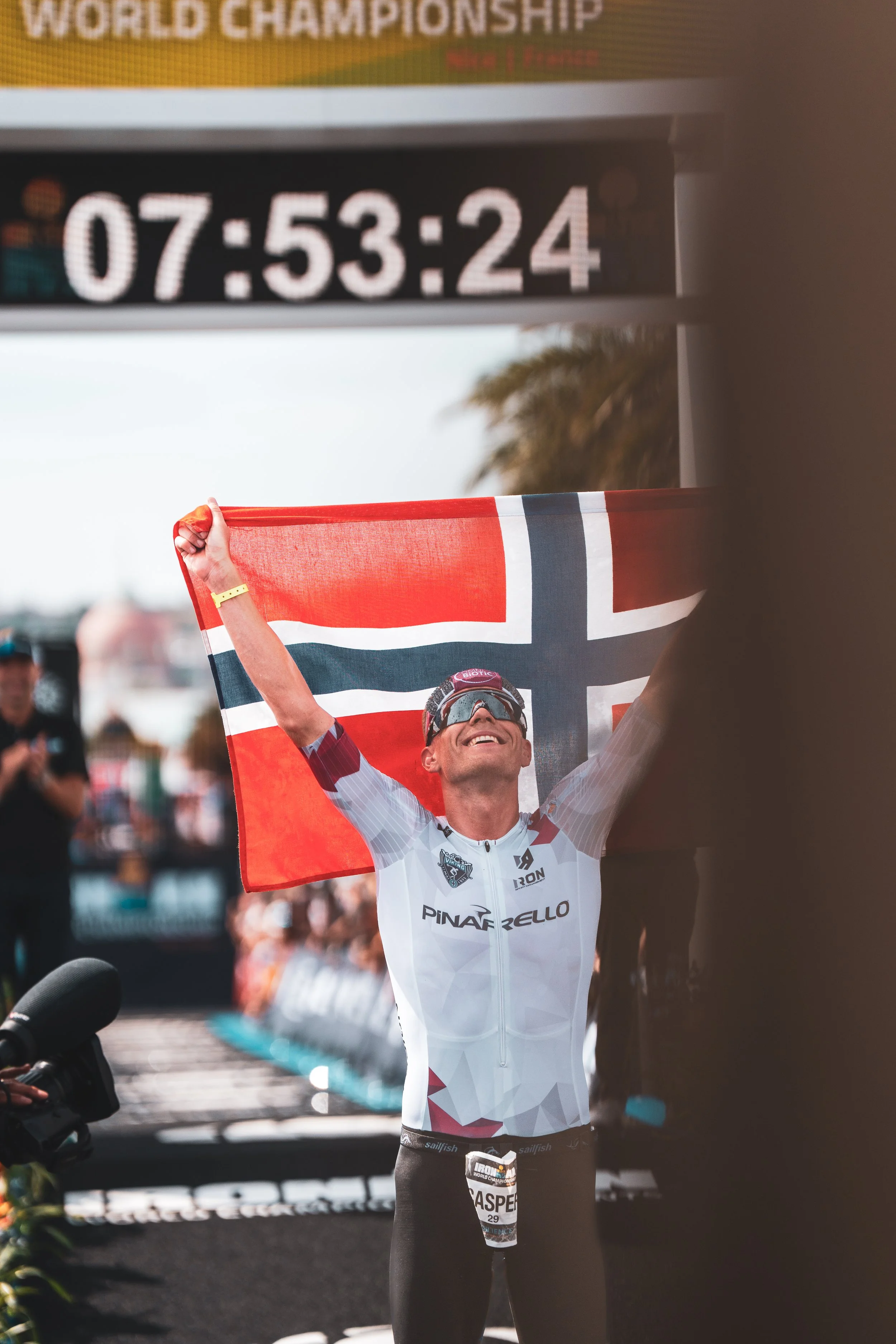Un athlète souriant, levant les bras avec un drapeau norvégien, célébrant sa victoire à l'arrivée d'une compétition de triathlon.