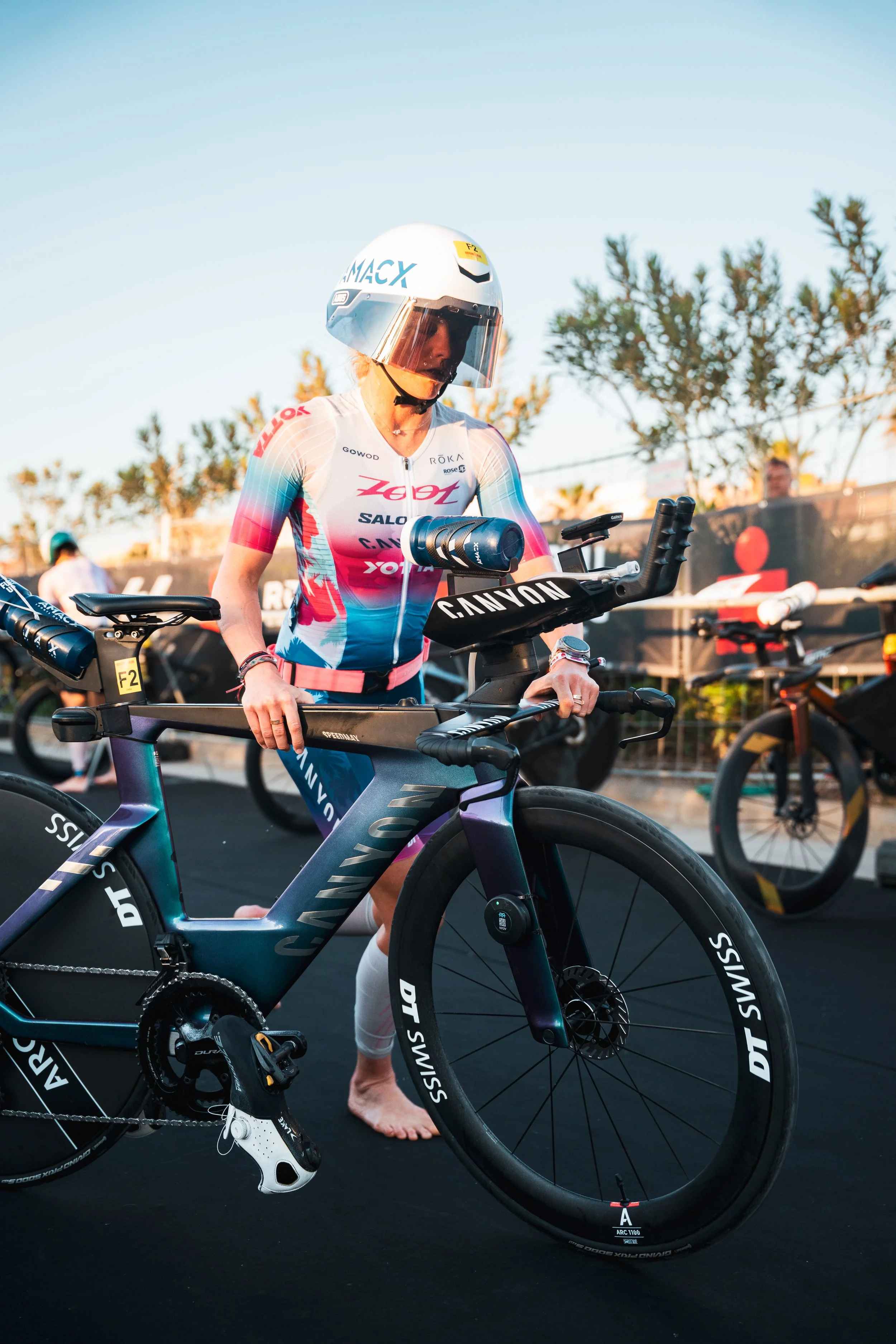 Une femme vêtue d'une maillot de cyclisme et portant un casque, se préparant avec un vélo de course de marque Canyon, dans un environnement extérieur avec d'autres vélos en arrière-plan.