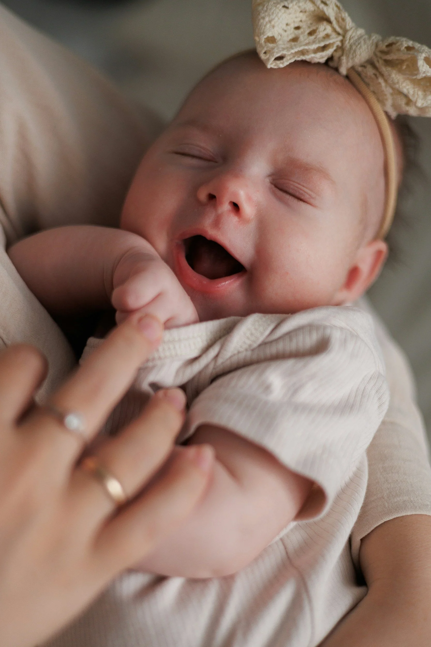Close-up of a yawning baby girl with a beige bow headband, being held by someone, possibly her mother, with a hand showing a wedding ring, touching her arm.