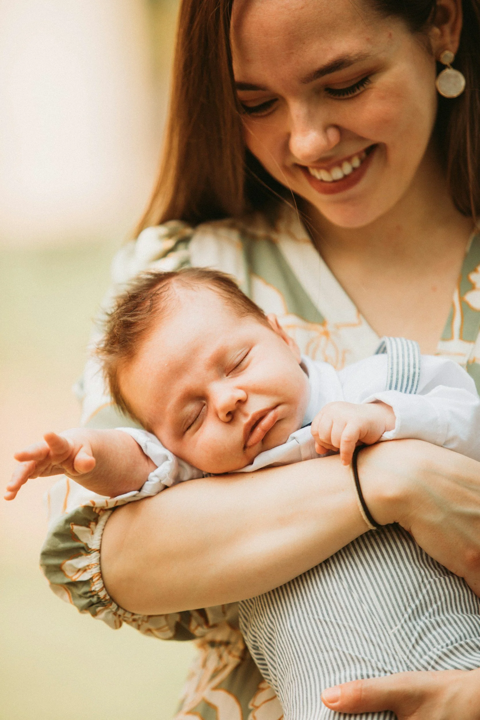 A woman holding a sleeping baby on her arm, smiling. The baby has closed eyes and is resting comfortably.