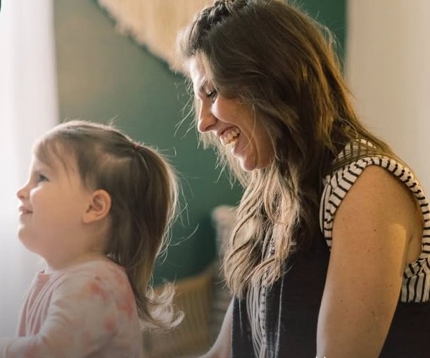 A woman and a young girl smiling and looking at each other indoors.