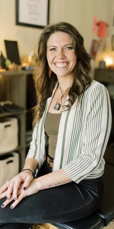 A woman with long wavy brown hair smiling, sitting in a cozy, well-lit room that has framed pictures and various decorative items in the background.