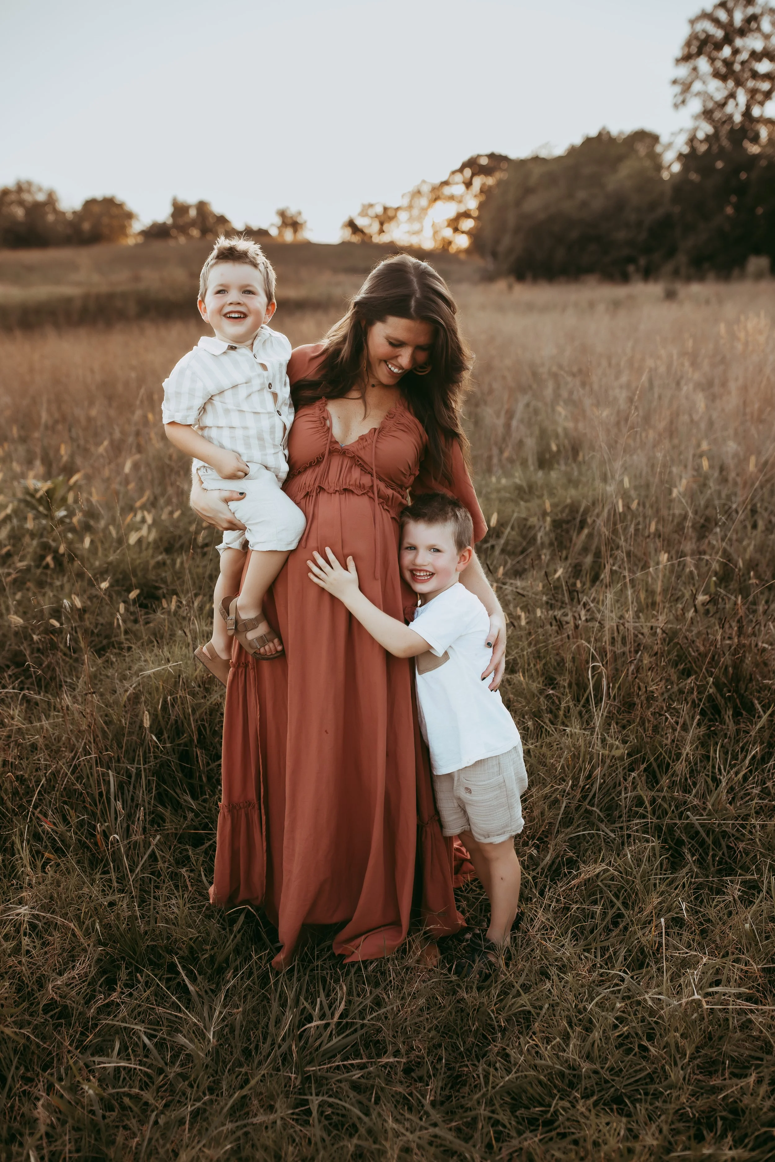 A woman with two young boys in a field during sunset, one boy sitting on her shoulder and hugging her, the other standing and hugging her waist, all smiling.