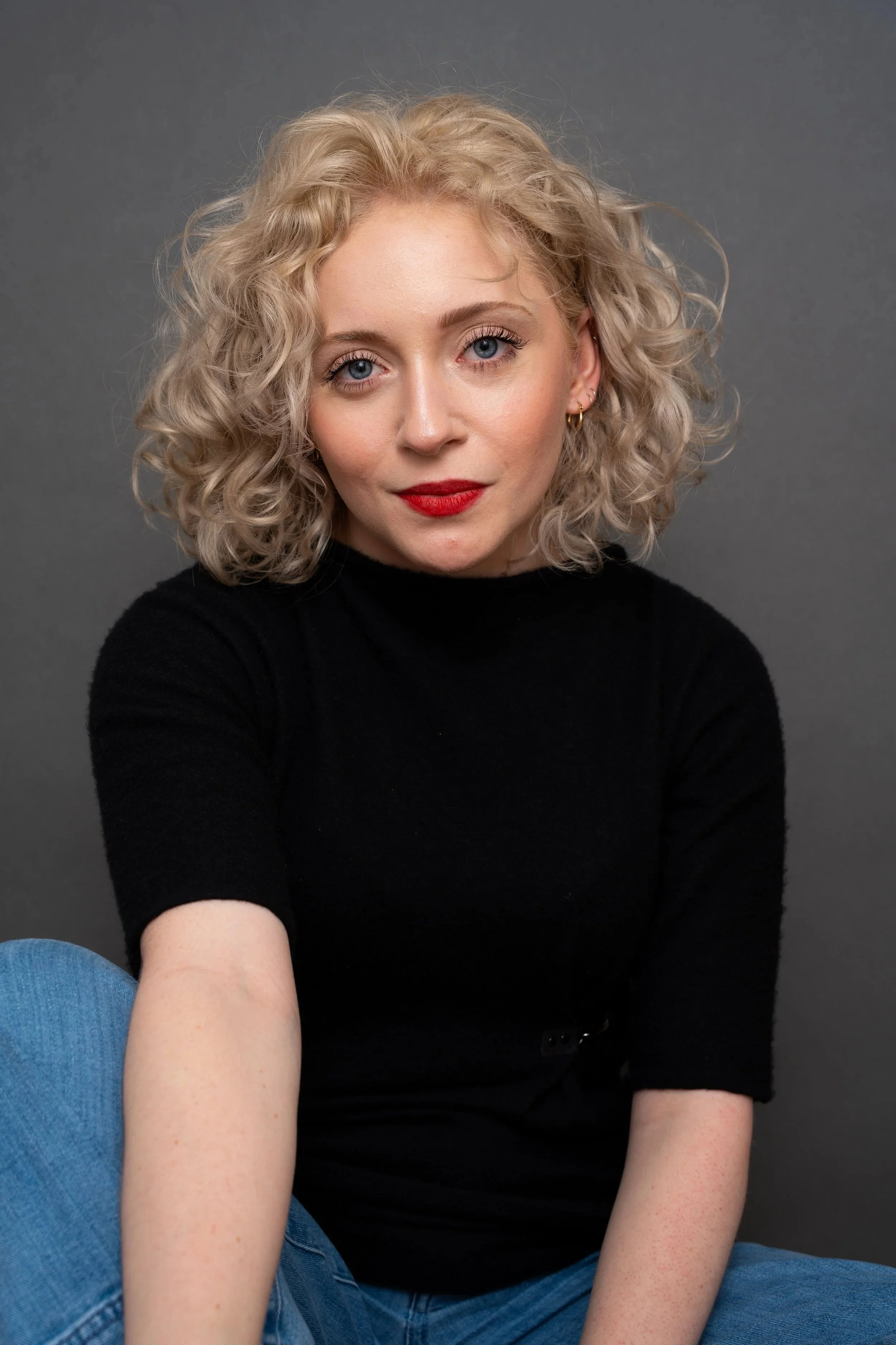 Photo of actress Mara Jade Koenig with blonde curly hair, wearing a black shirt and jeans sitting with one leg up.