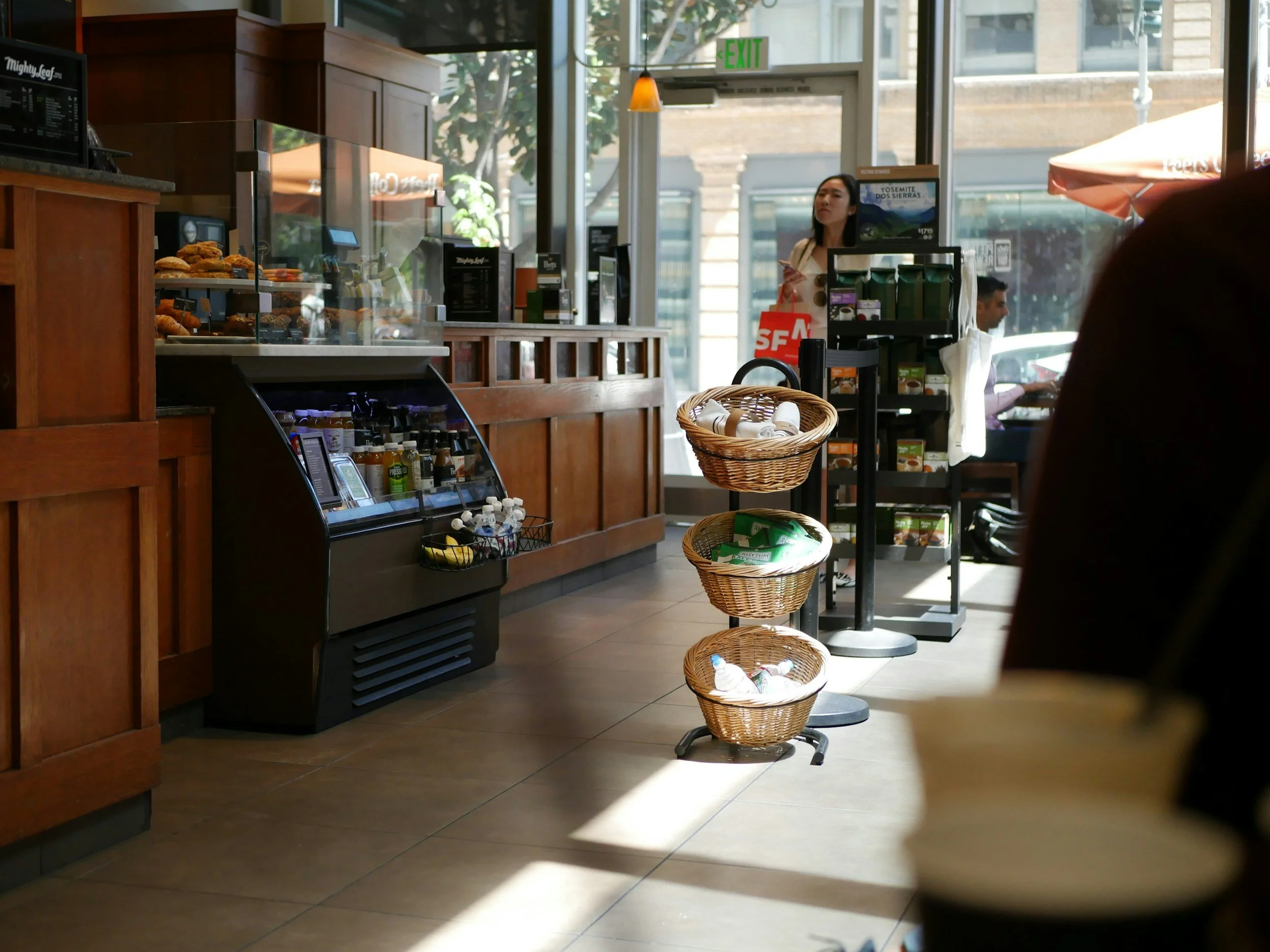 Interior of a coffee shop with display case of baked goods, refrigerated section, shelves with bottled drinks, and tables near large windows with sunlight and outdoor seating.