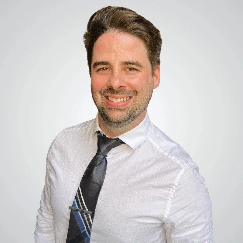 A man with short dark hair and a beard, smiling, wearing a white dress shirt and a dark striped tie, against a plain white background.