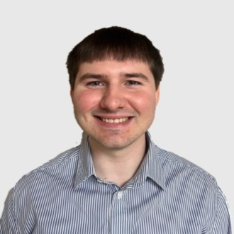 Portrait of a young man with short brown hair, smiling, wearing a blue striped button-up shirt, against a plain light gray background.