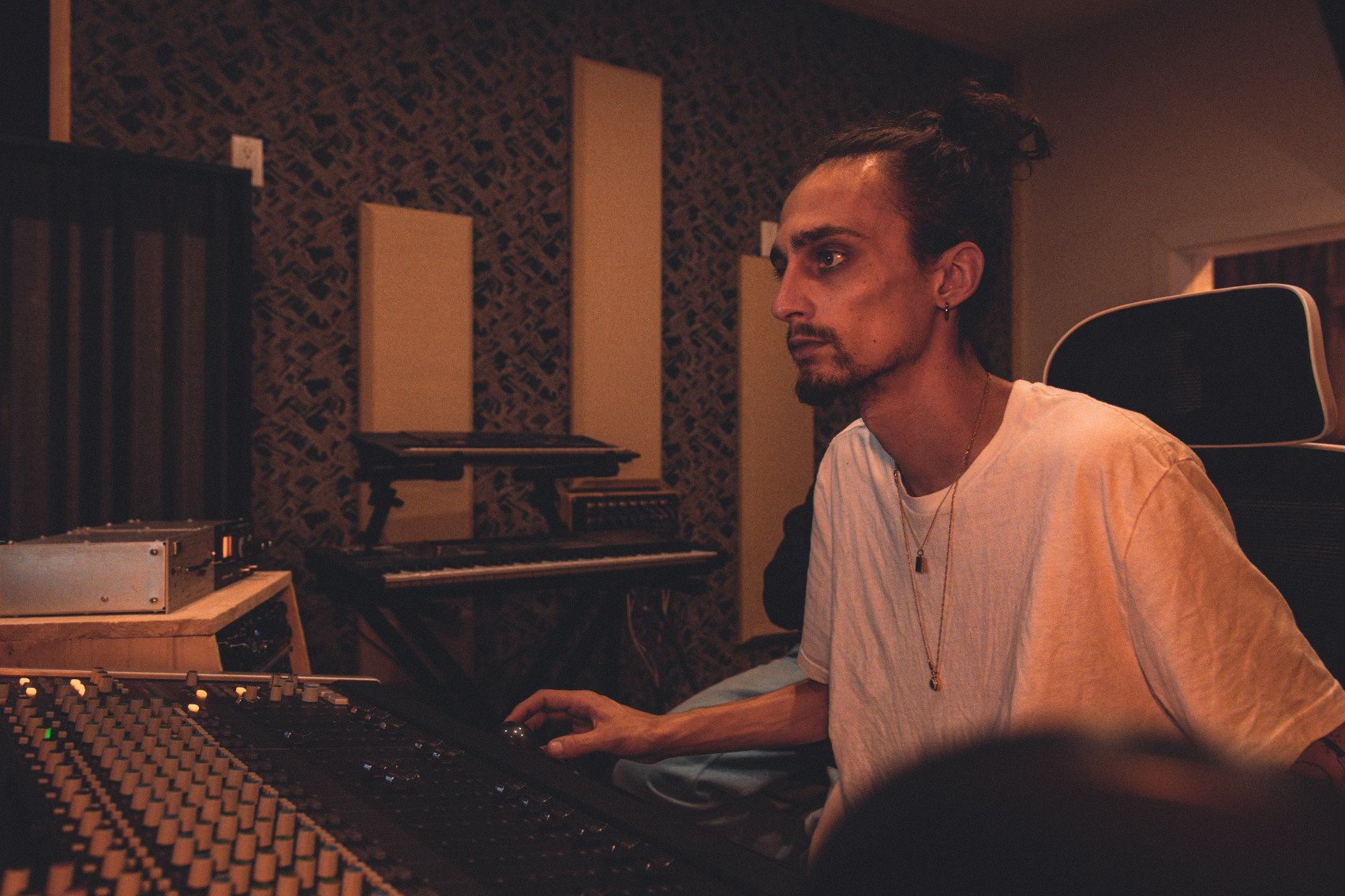 A person with long dark hair tied back, wearing a white t-shirt and jewelry, sitting at a music mixing console in a recording studio, surrounded by sound equipment and acoustic panels.
