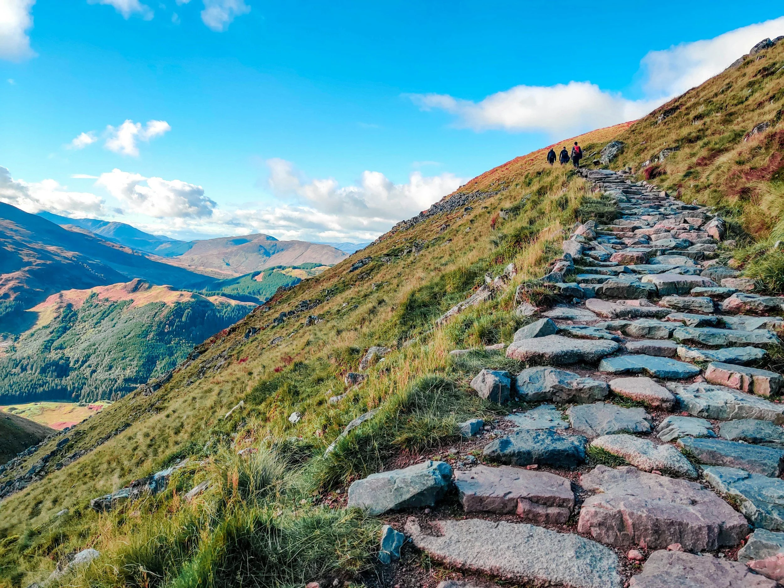 Three people hiking on a rocky trail up a grassy hillside with mountains and a blue sky in the background.